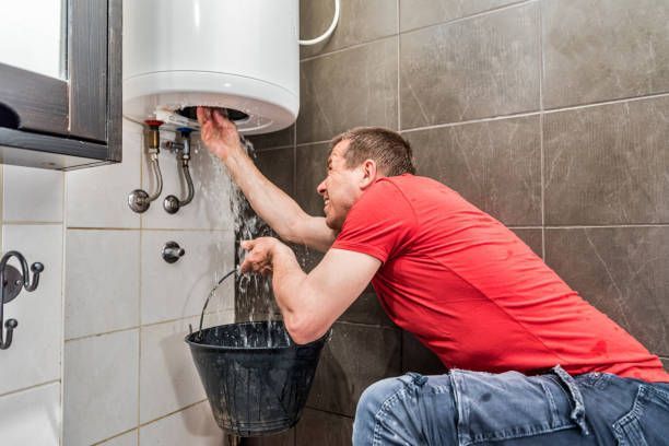 A person in a red shirt holding a bucket under a leaking water heater mounted on a tiled wall.