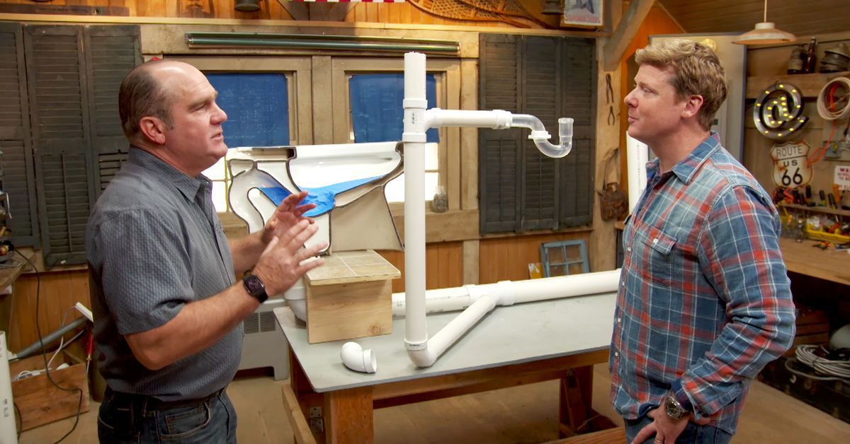 Two people in a workshop discuss a plumbing display featuring white PVC pipes and a clear P-trap on a wooden table.