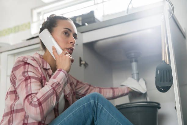 A person talks on a phone while using a cloth to wipe underneath a sink into a bucket.