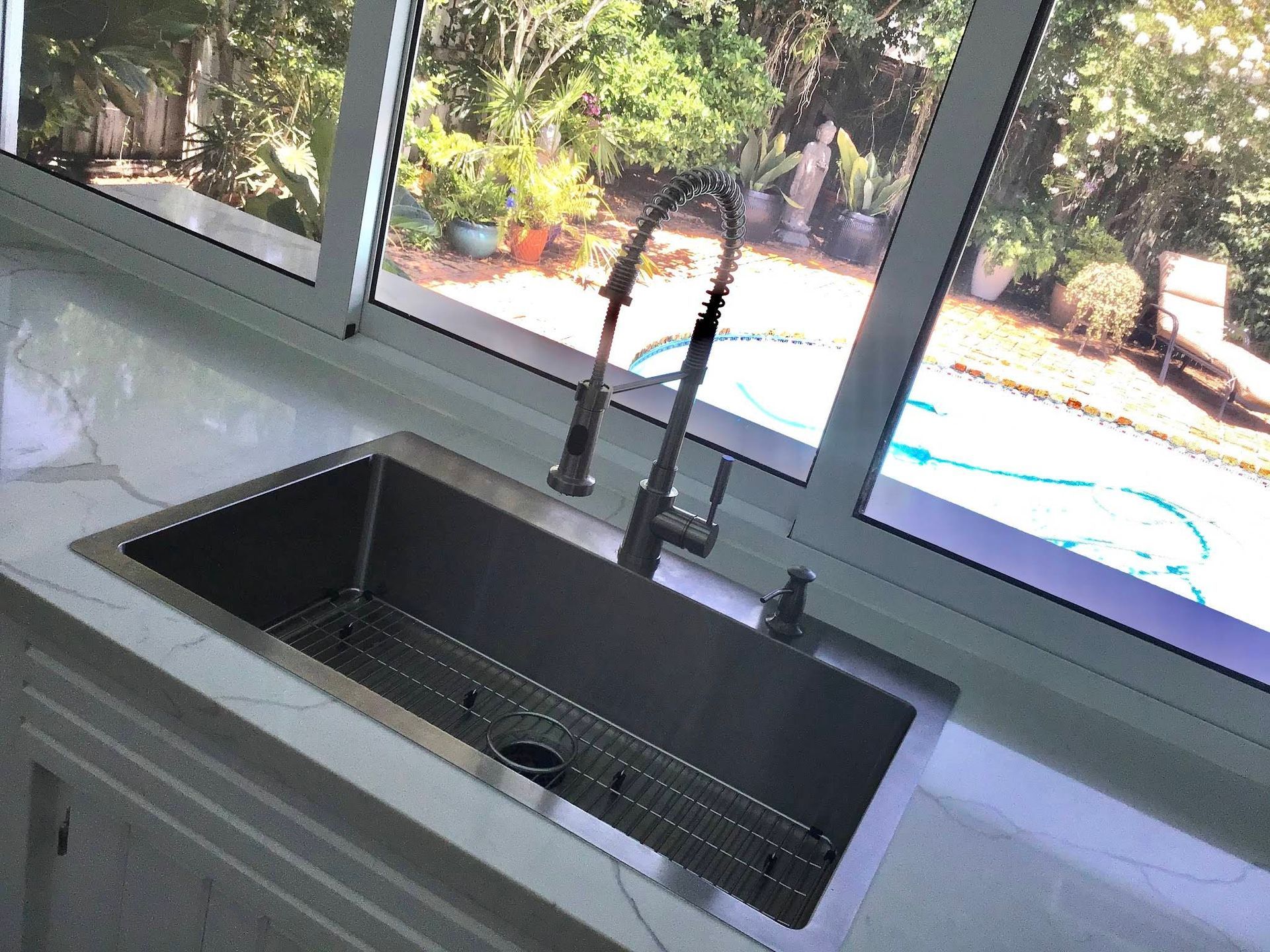 A stainless steel kitchen sink with a tall gooseneck faucet, set into a light-colored countertop before a large window.
