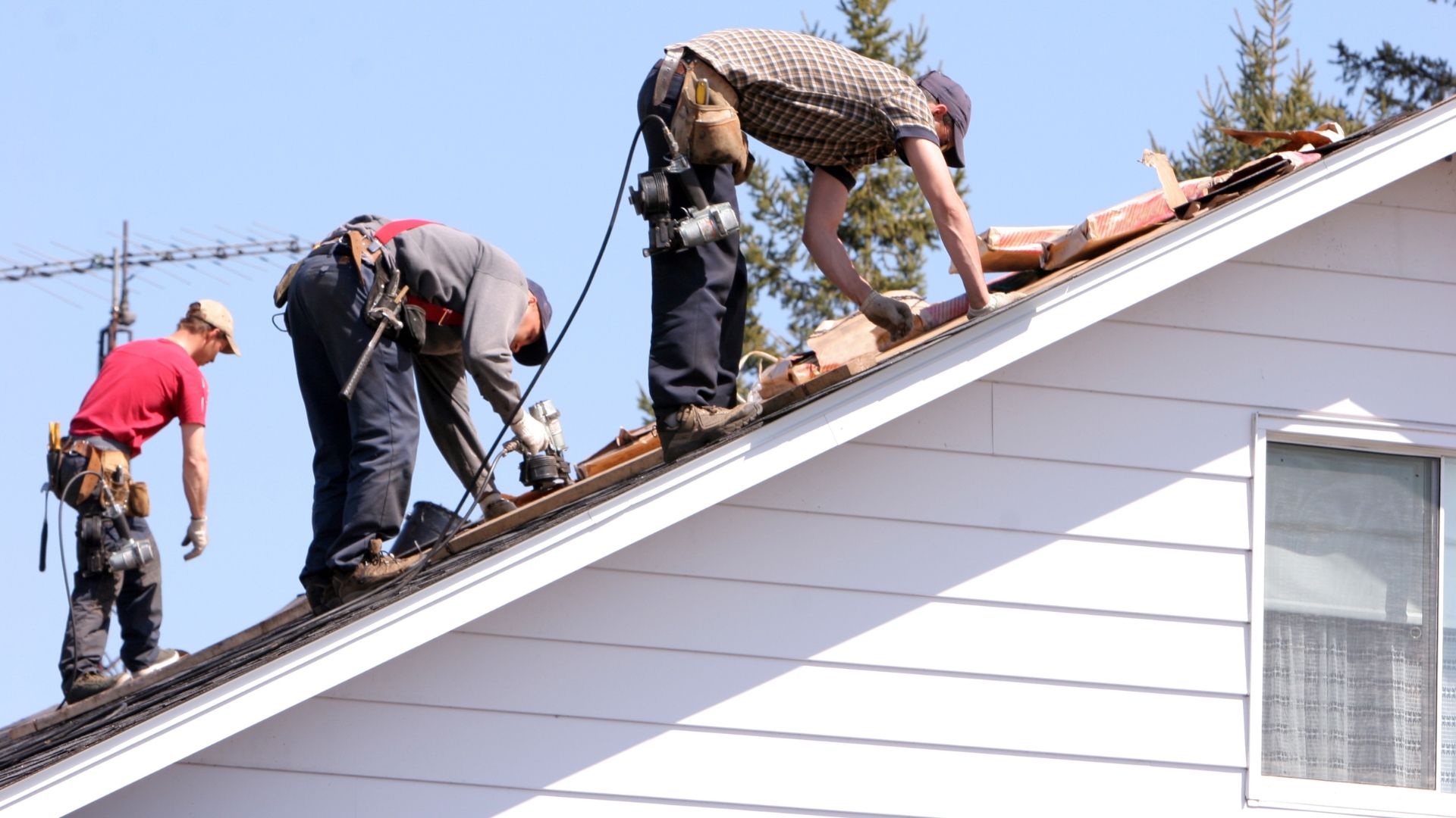 Three roofers on a rooftop installing tiles. Sunny day, white house, blue sky.