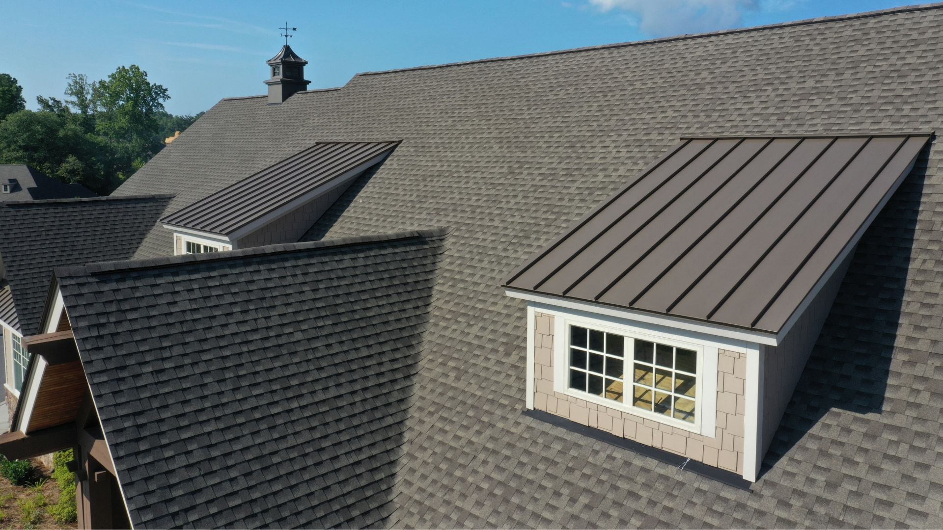 Brown shingled roof with multiple dormers, one with a metal roof. A weather vane is atop the roof.