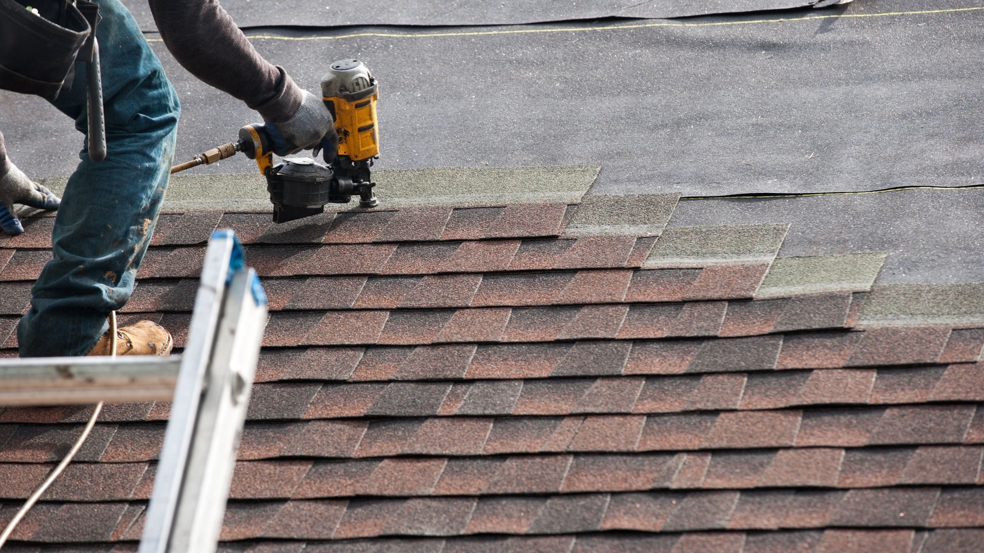 Person using a nail gun to install roofing shingles on a rooftop.