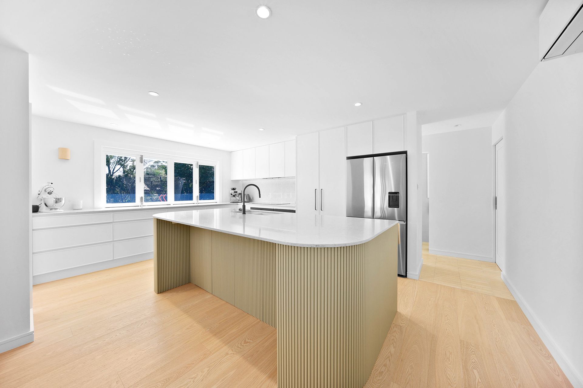 Modern white kitchen with light wood floors, a gold fluted island, and stainless steel refrigerator — Above & Beyond Interiors Custom Joinery in Bellambi, NSW