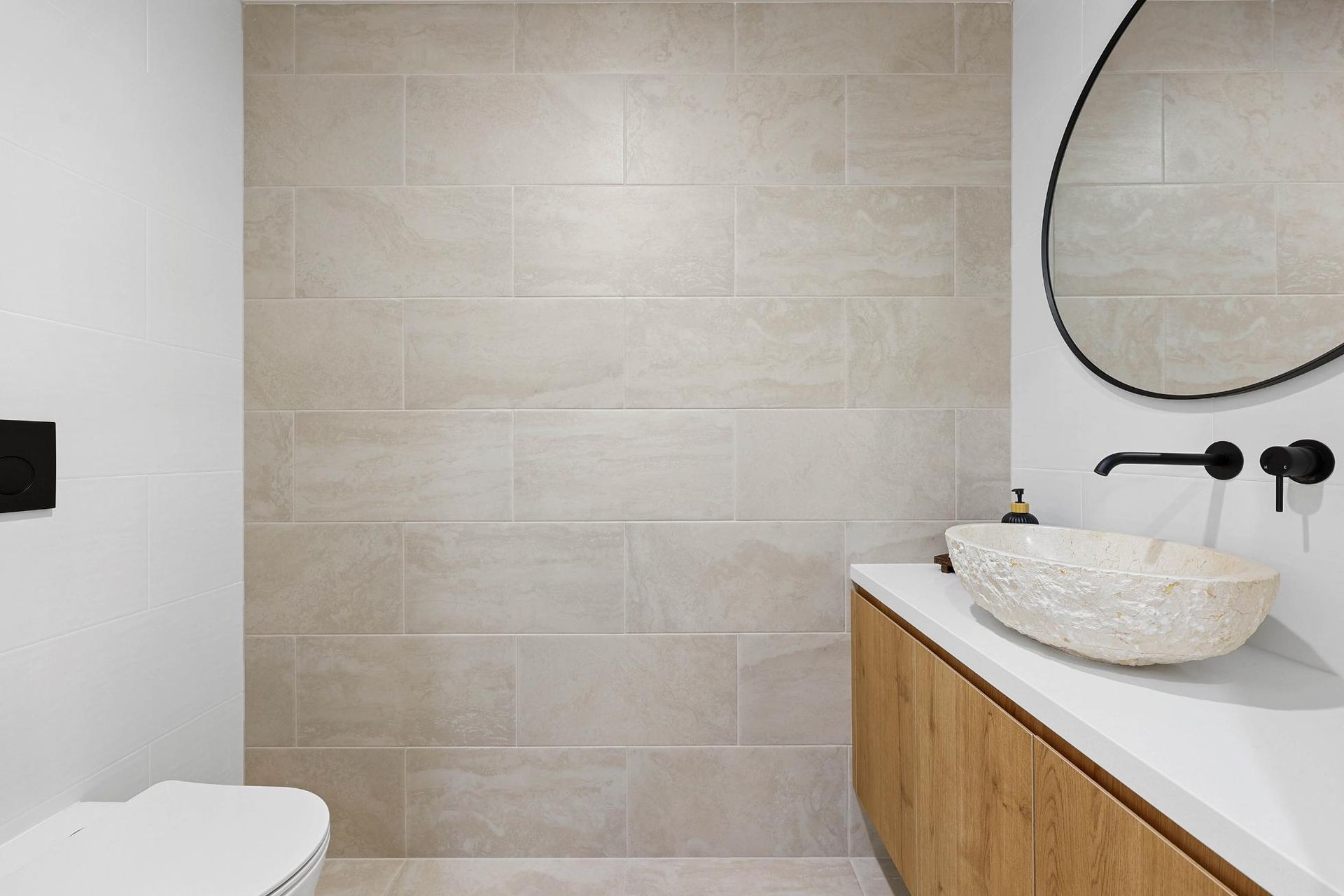 A Bathroom with Light Tan Tiled Wall, Wooden Vanity, White Countertop, Stone Sink, Black Fixtures, and Round Mirror — Above & Beyond Interiors Custom Joinery in Abbortsbury, NSW