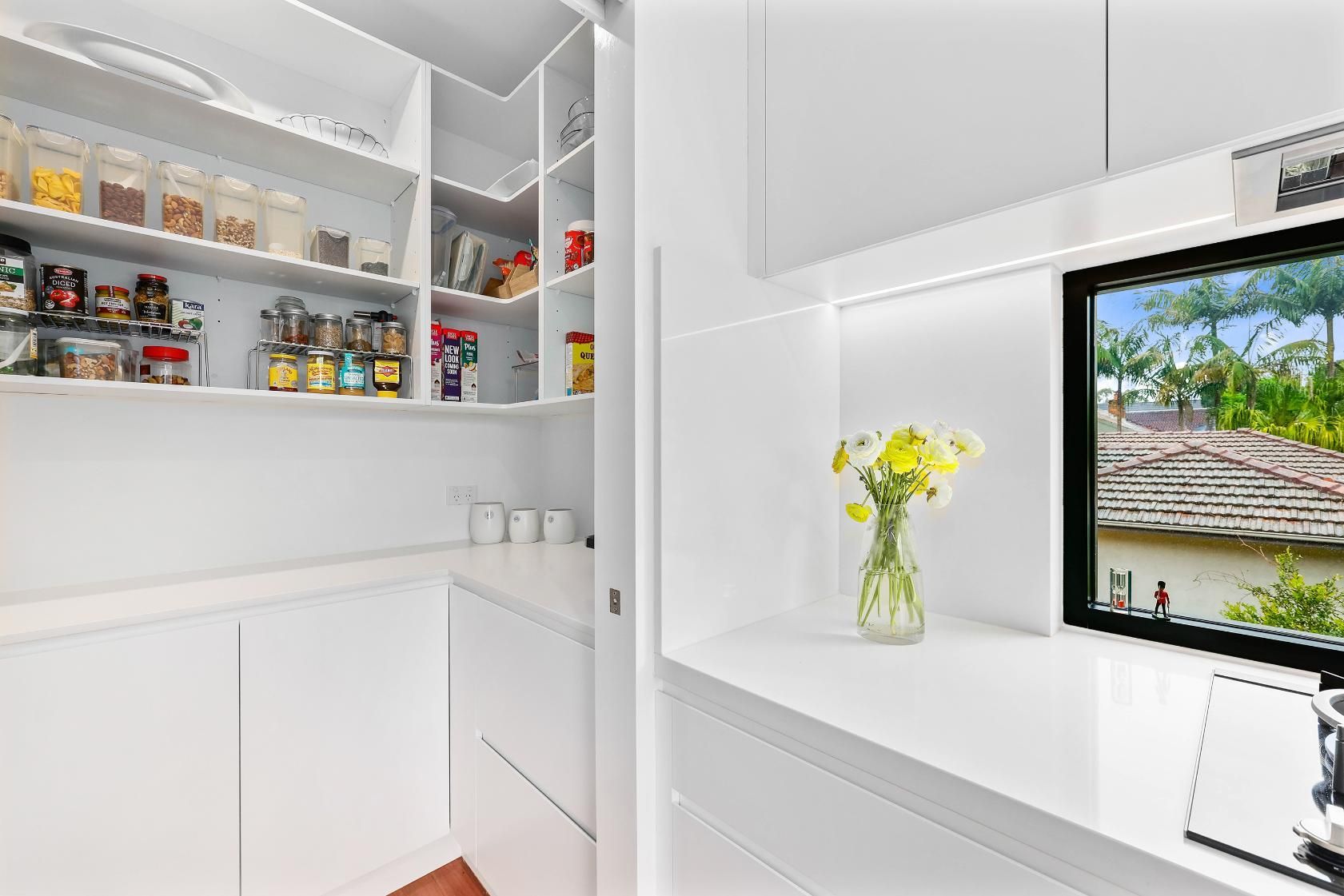 White Pantry with Shelves of Food Items, and Built-In Desk with Flower Vase, Next to Window — Above & Beyond Interiors Custom Joinery in Killara, NSW