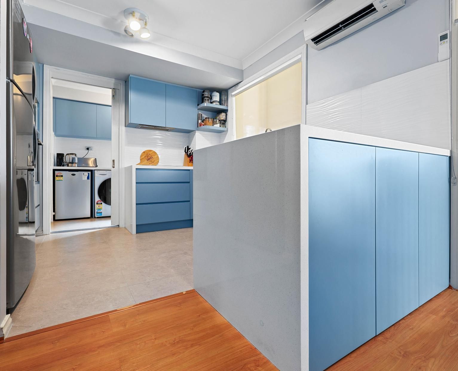 Blue and White Kitchen with Cabinetry and Island. Laundry Machines Visible in The Background — Above & Beyond Interiors Custom Joinery in Macquarie Fields, NSW