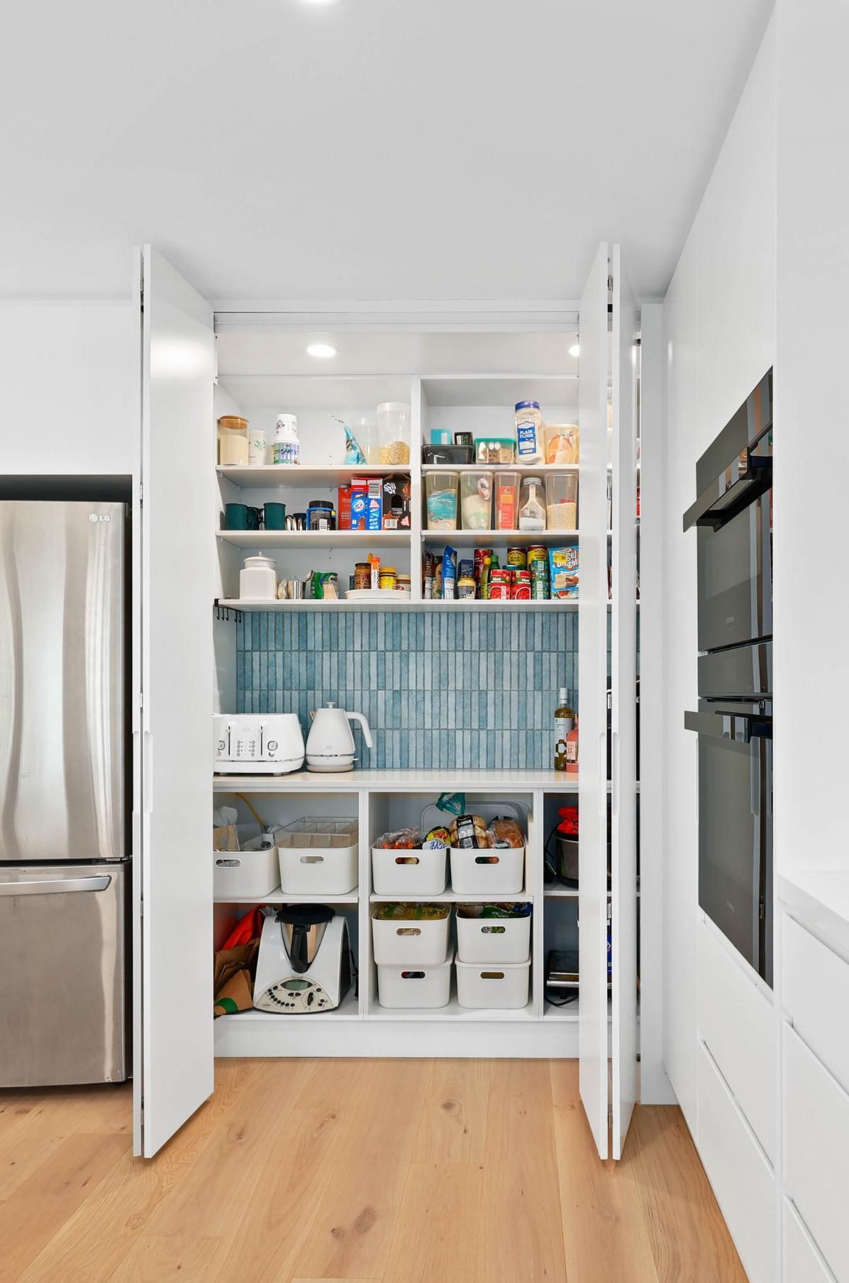 White Pantry with Open Doors, Displaying Organized Shelves of Food Items; Next to A Refrigerator and Oven — Above & Beyond Interiors Custom Joinery in North Curl Curl, NSW