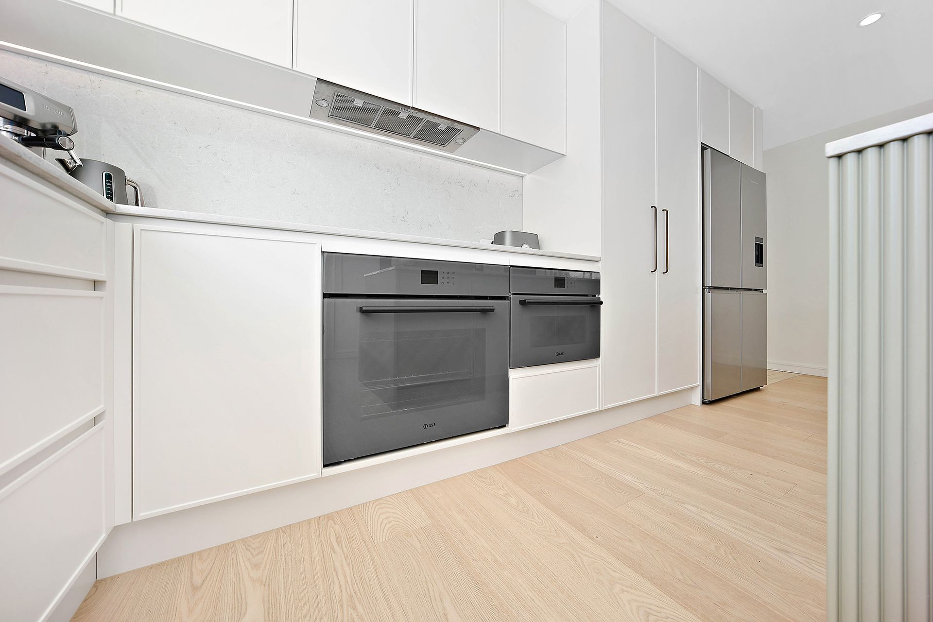 Modern white kitchen with light wood floors, built-in ovens, and stainless steel refrigerator — Above & Beyond Interiors Custom Joinery in Bellambi, NSW