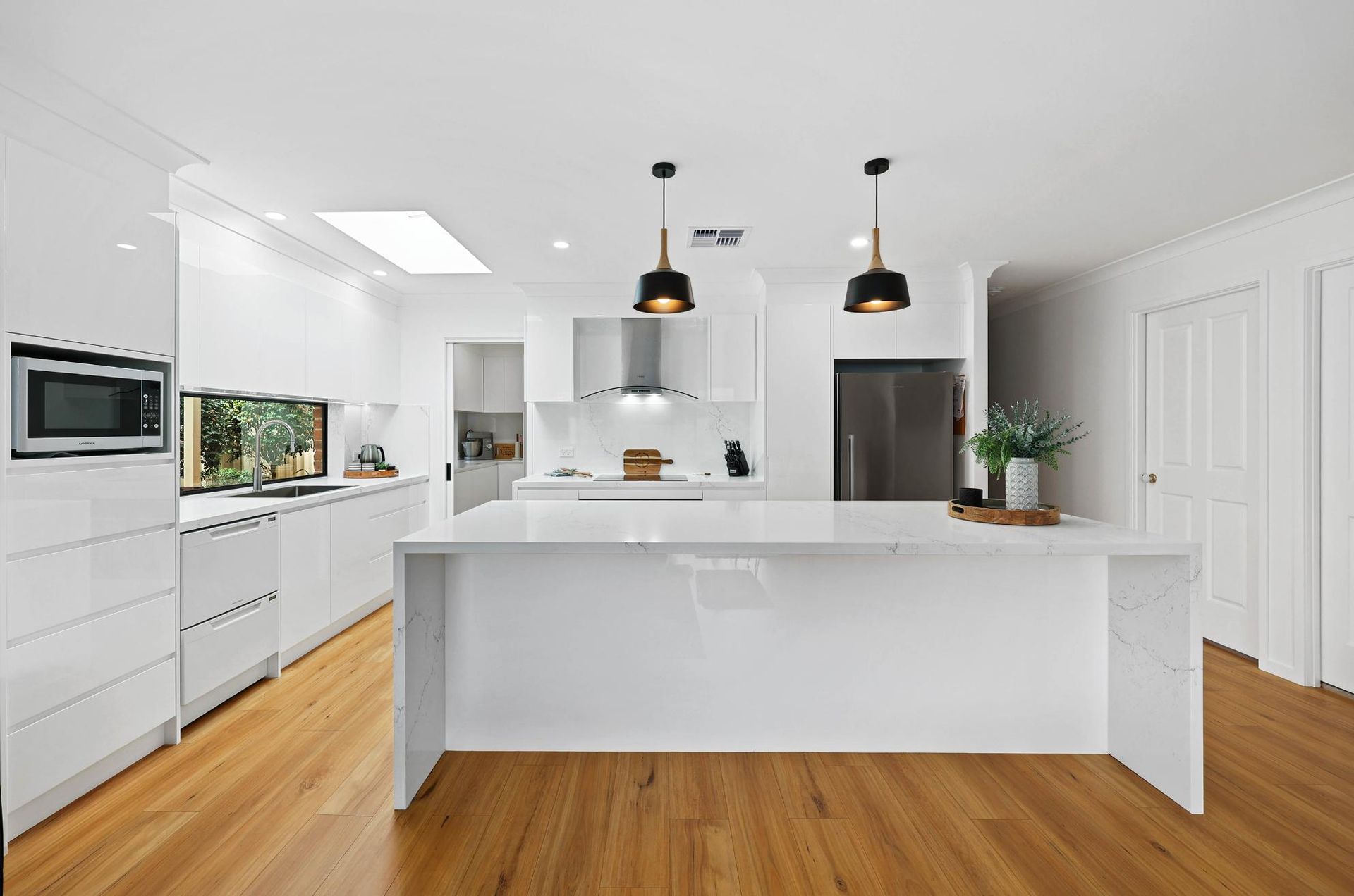 Modern White Kitchen with Island, Wood Floors, Black Pendant Lights, and Stainless Steel Appliances — Above & Beyond Interiors Custom Joinery in Wattle Grove, NSW