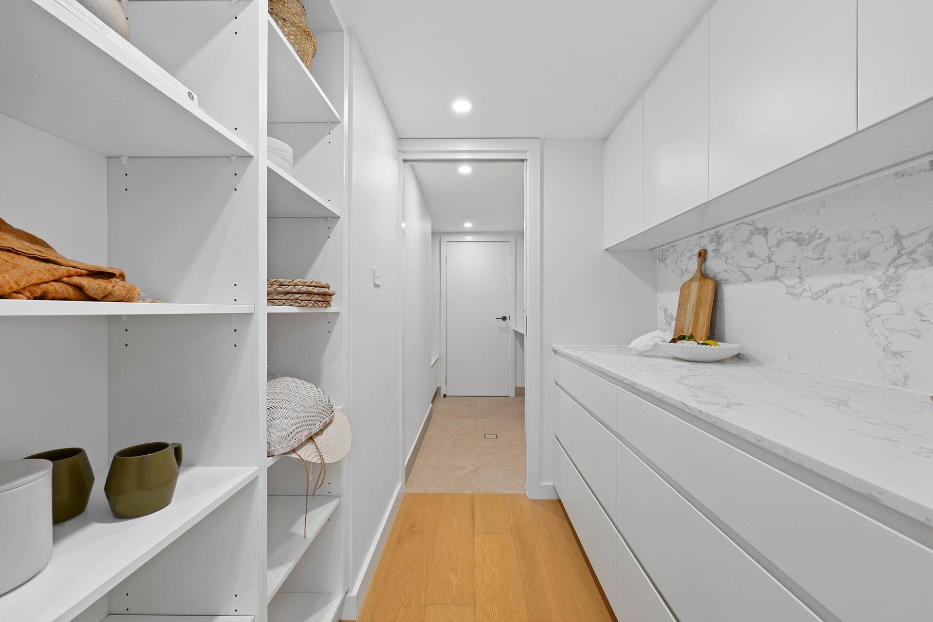 White Pantry with Shelves on One Side, Counter and Cabinets on The Other, a Door at The End — Above & Beyond Interiors Custom Joinery in Thirroul, NSW