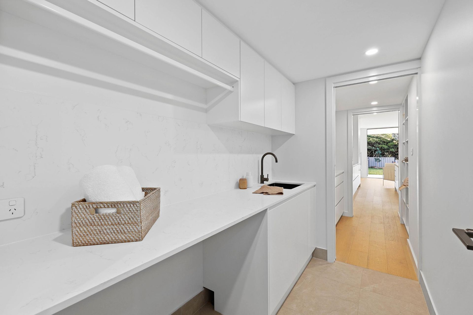 White Laundry Room with Cabinets, Countertop, Black Faucet, Wicker Basket, and Doorway — Above & Beyond Interiors Custom Joinery in Thirroul, NSW