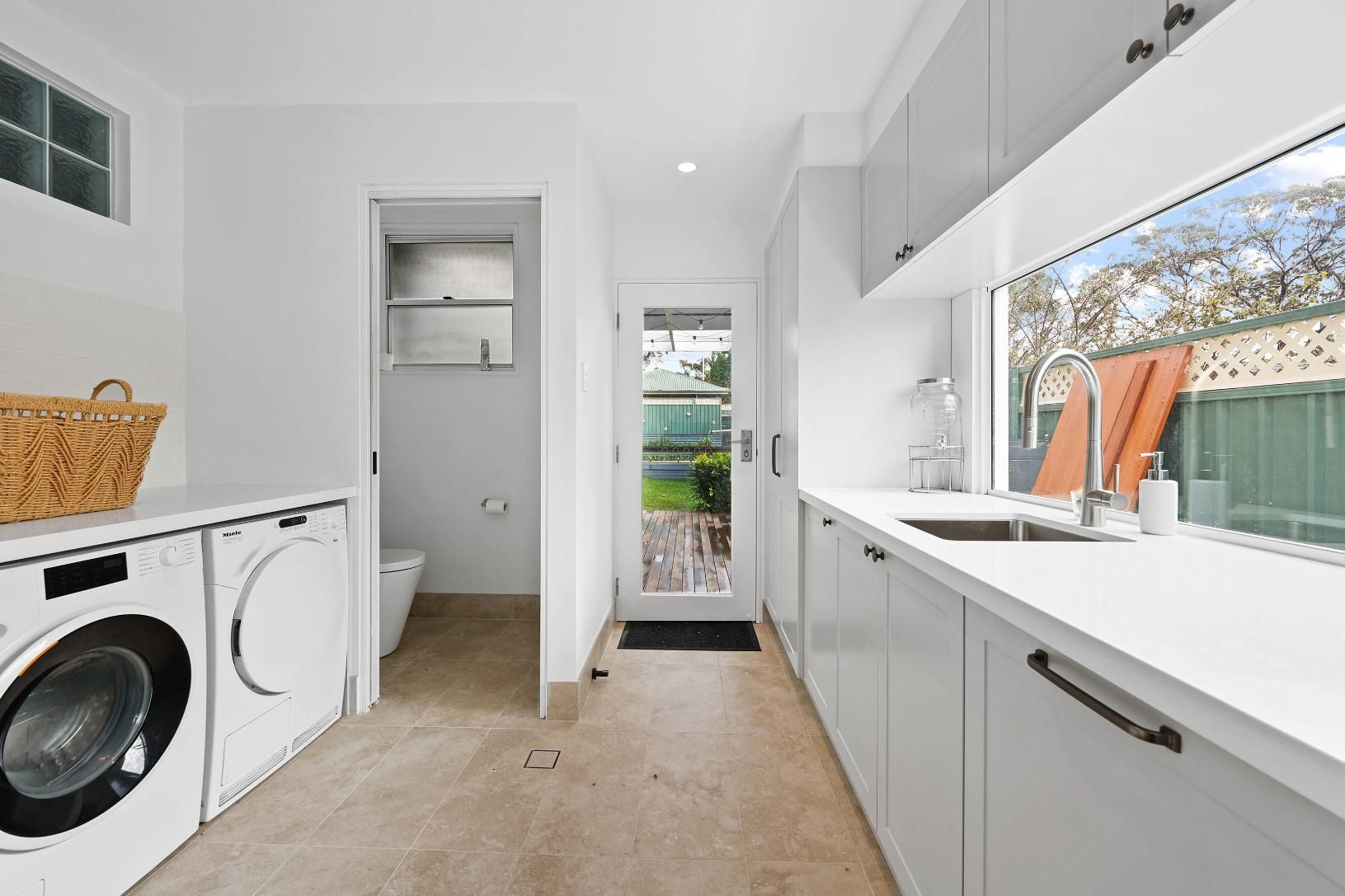 Laundry Room with White Cabinets, Washer/dryer, Sink, and A View of A Green Yard — Above & Beyond Interiors Custom Joinery in Yowie Bay, NSW