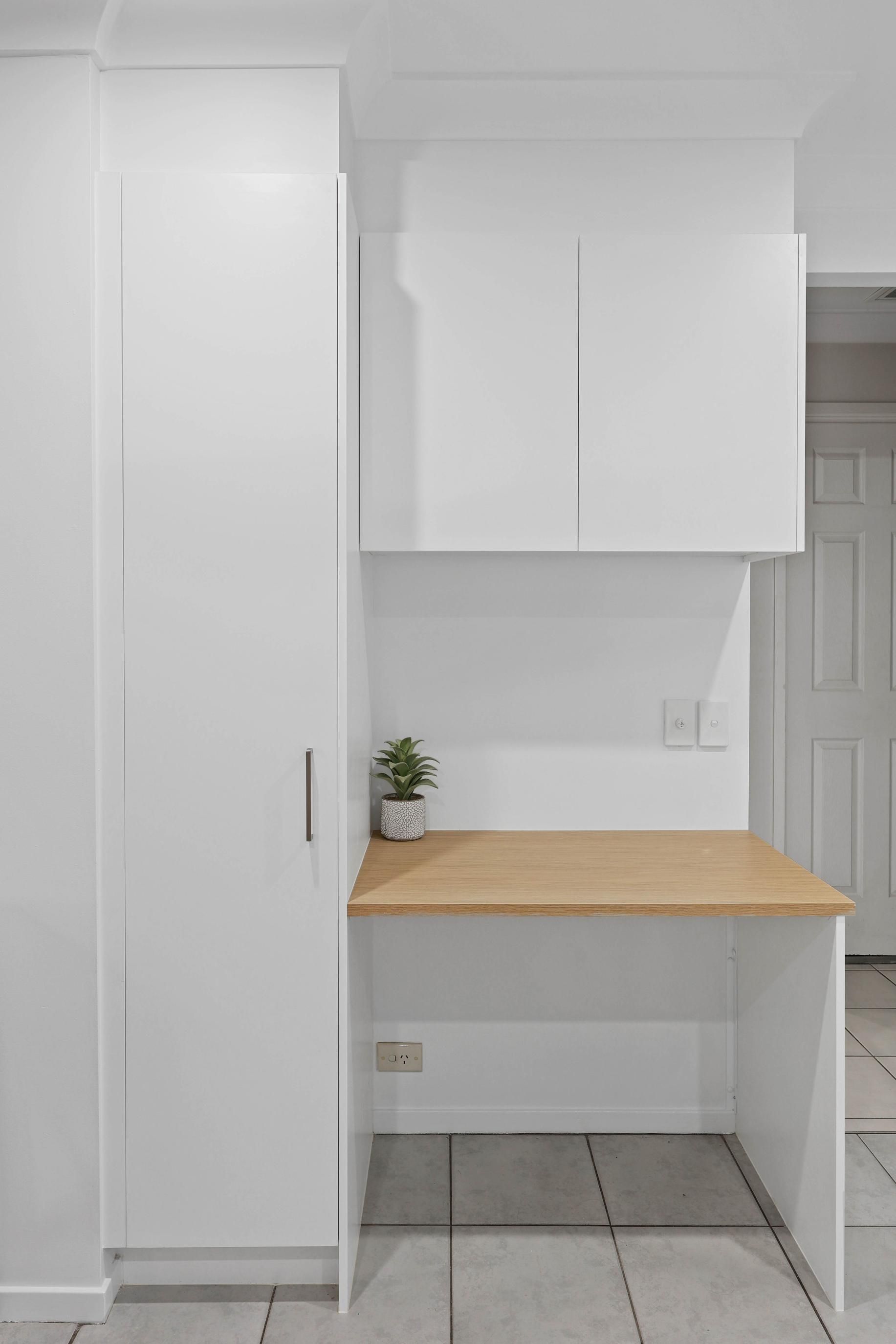White Built-In Desk with Wood-Tone Countertop, Overhead Cabinets, and Tall Cabinet. a Small Plant Sits on The Desk — Above & Beyond Interiors Custom Joinery in Currans Hill, NSW