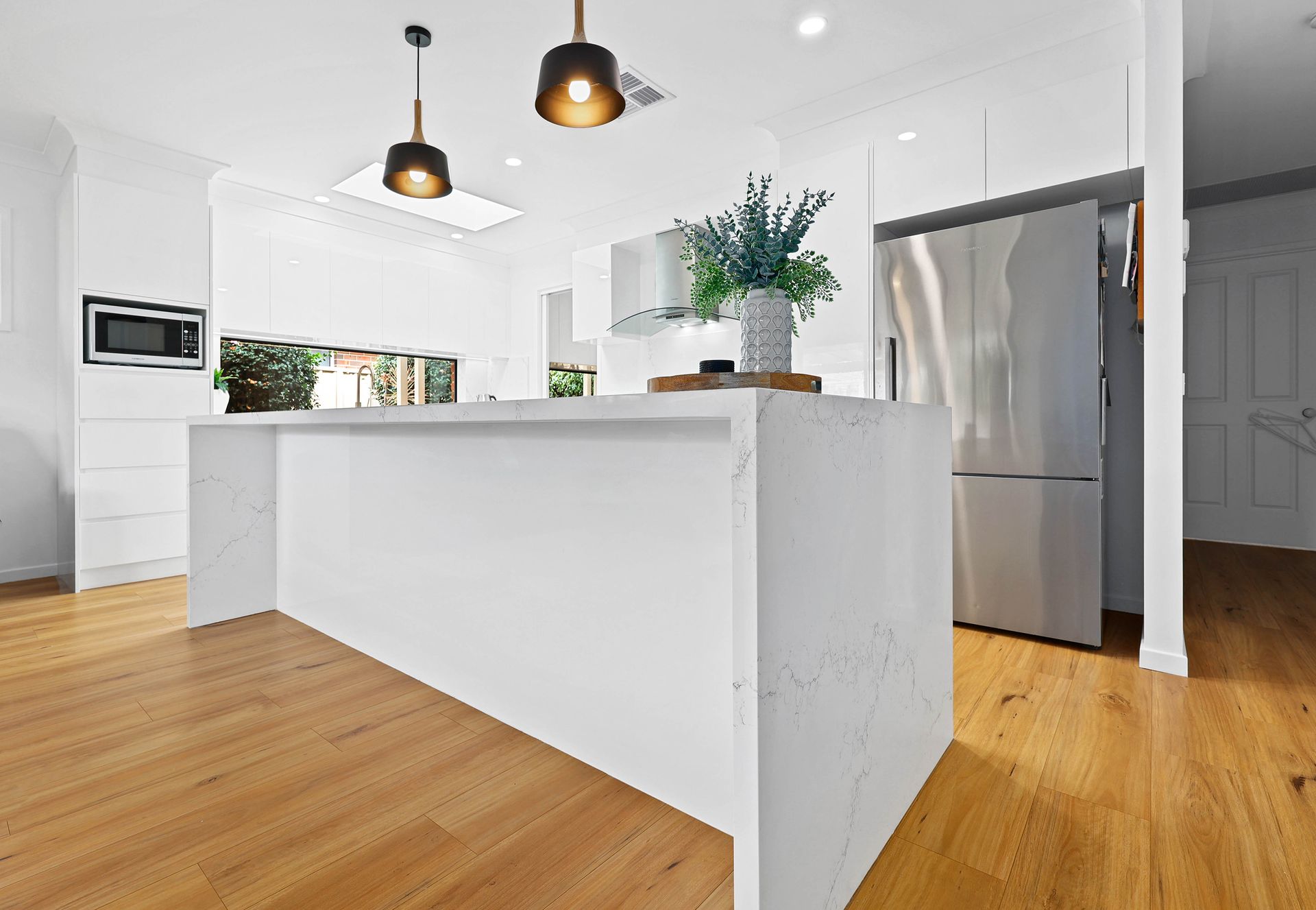 Modern white kitchen with island, wooden floors, stainless steel fridge, and pendant lights — Above & Beyond Interiors in Smeaton Grange, NSW