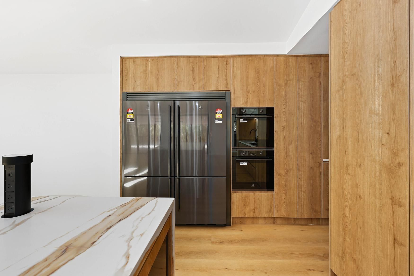 Modern Kitchen with Wood-Grain Cabinetry, Stainless Steel Refrigerator, and Marble Countertop Island — Above & Beyond Interiors Custom Joinery in Abbortsbury, NSW