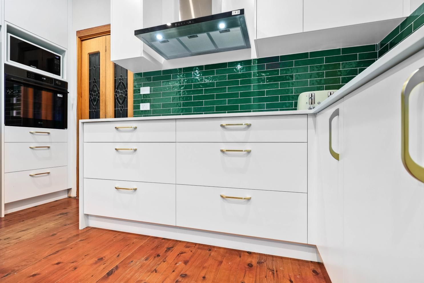 White Kitchen with Gold Hardware, Green Tiled Backsplash, and Wood Flooring — Above & Beyond Interiors Custom Joinery in Minto, NSW