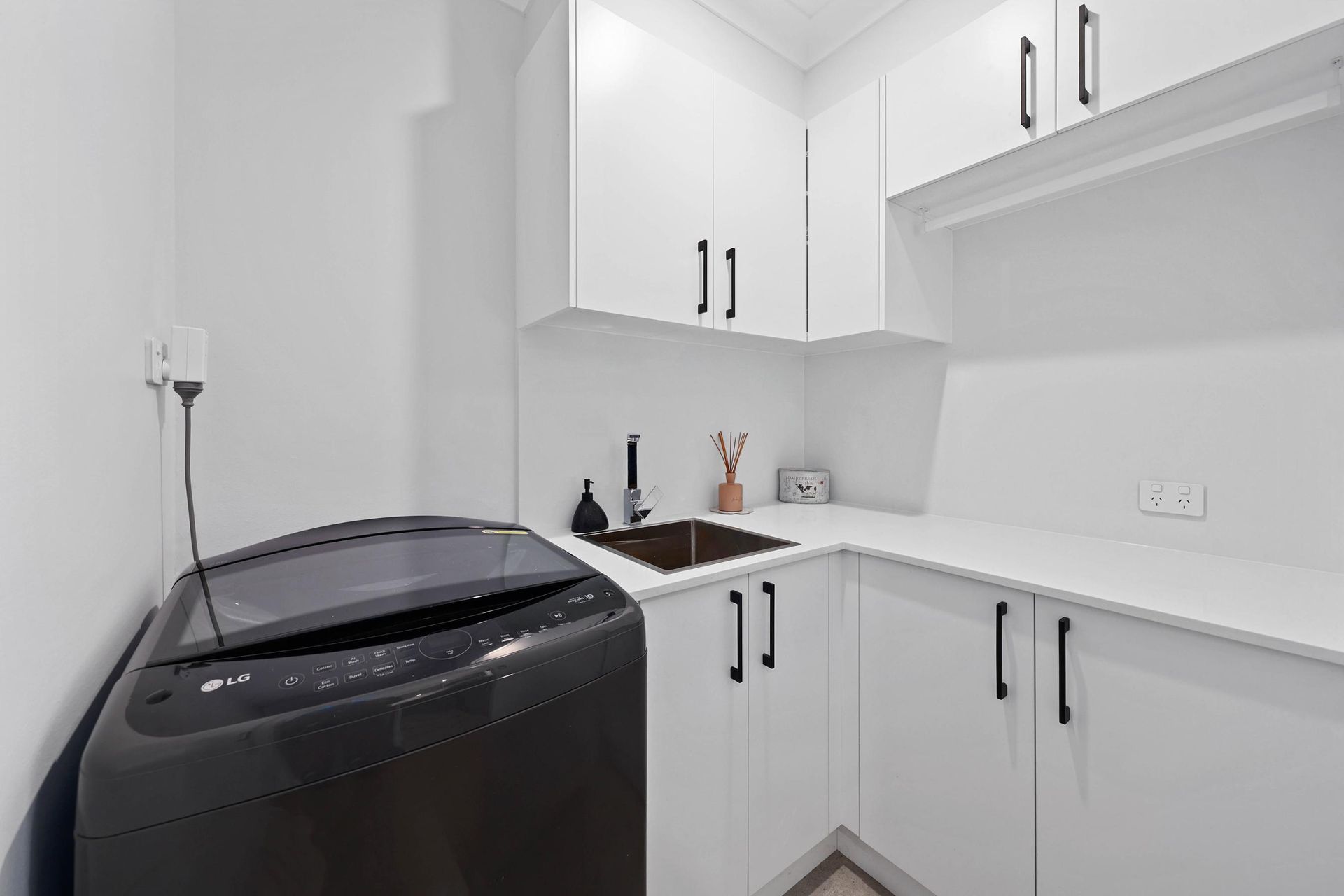 A Modern White Laundry Room with A Black Washing Machine, Sink, and Cabinets — Above & Beyond Interiors Custom Joinery in Bradbury, NSW