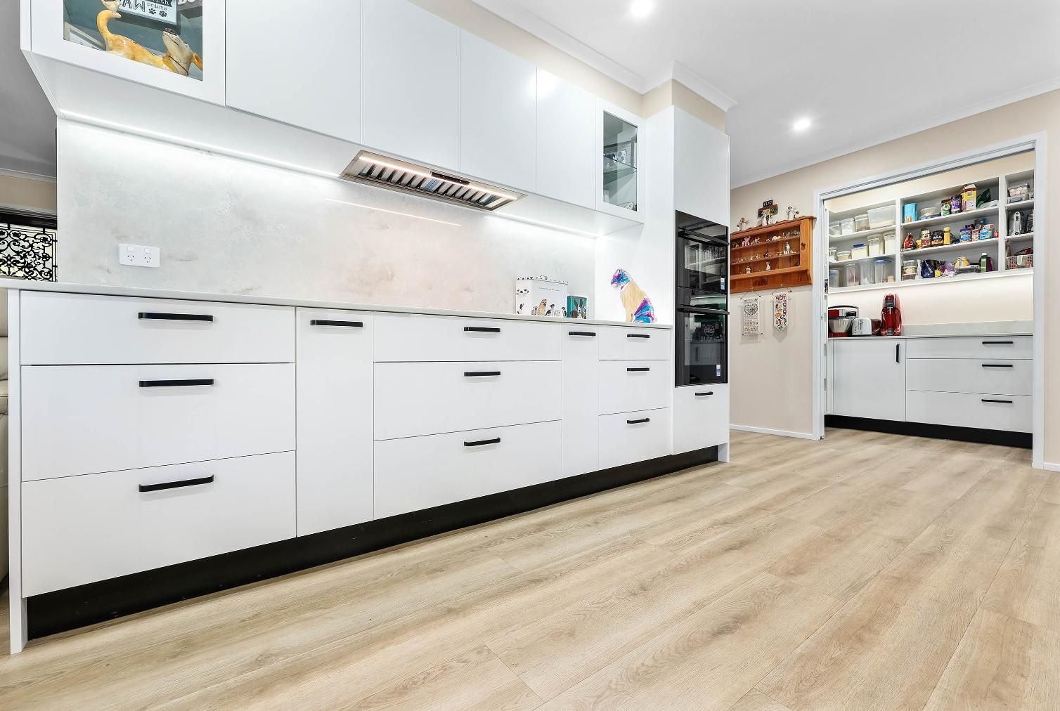 Modern White Kitchen with Light Wood Floor. Cabinets with Black Handles, Appliances, and A View Into Another Kitchen — Above & Beyond Interiors Custom Joinery in Bradbury, NSW