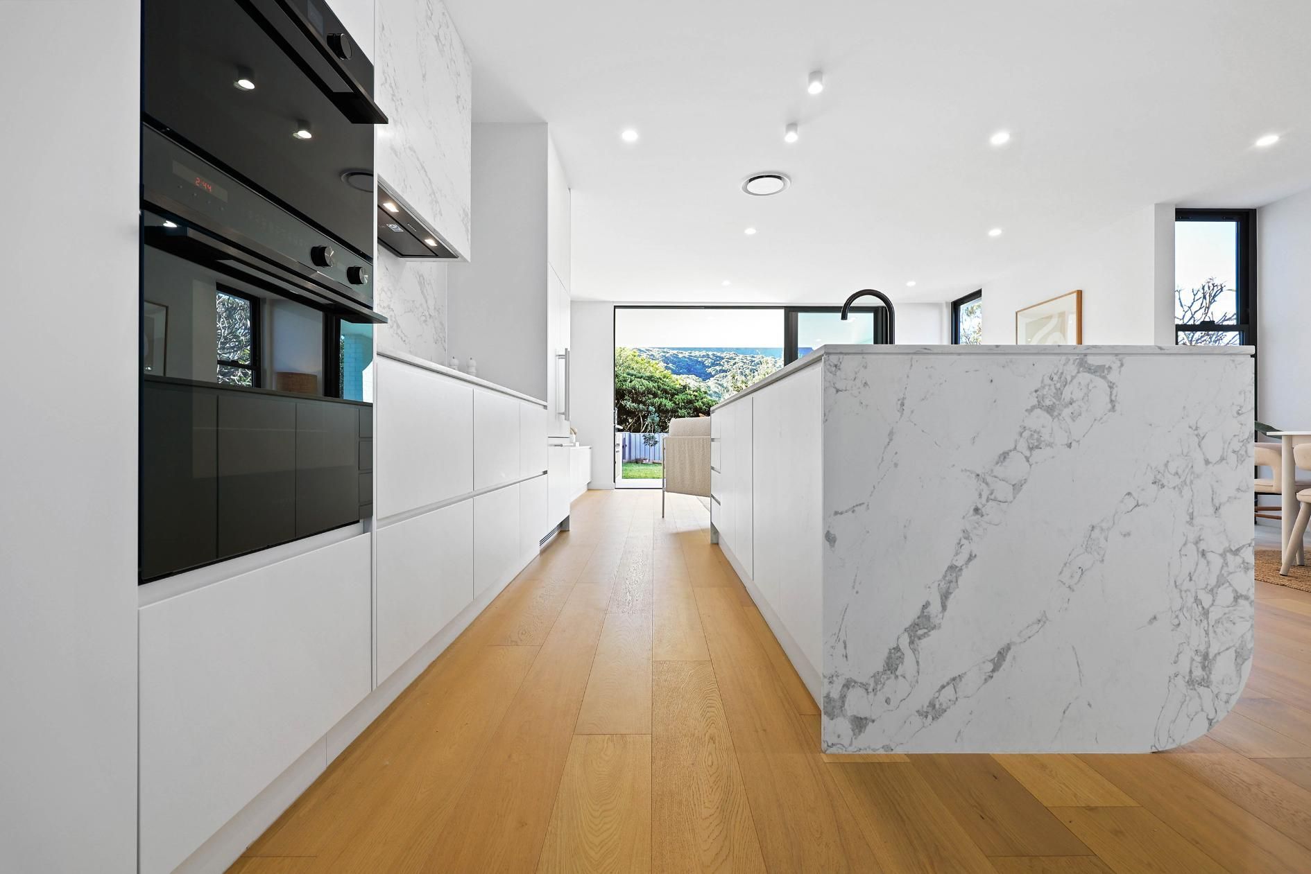 Modern White Kitchen with Island and Black Oven, Wooden Floors and A View to The Yard — Above & Beyond Interiors Custom Joinery in Thirroul, NSW