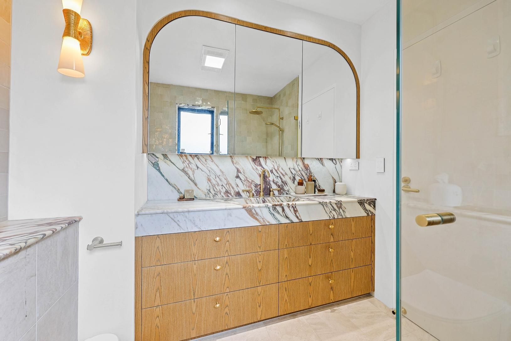 Bathroom with Wooden Vanity, Marble Countertop, Arched Mirror, Gold Accents, and Glass Shower — Above & Beyond Interiors Custom Joinery in Double Bay, NSW