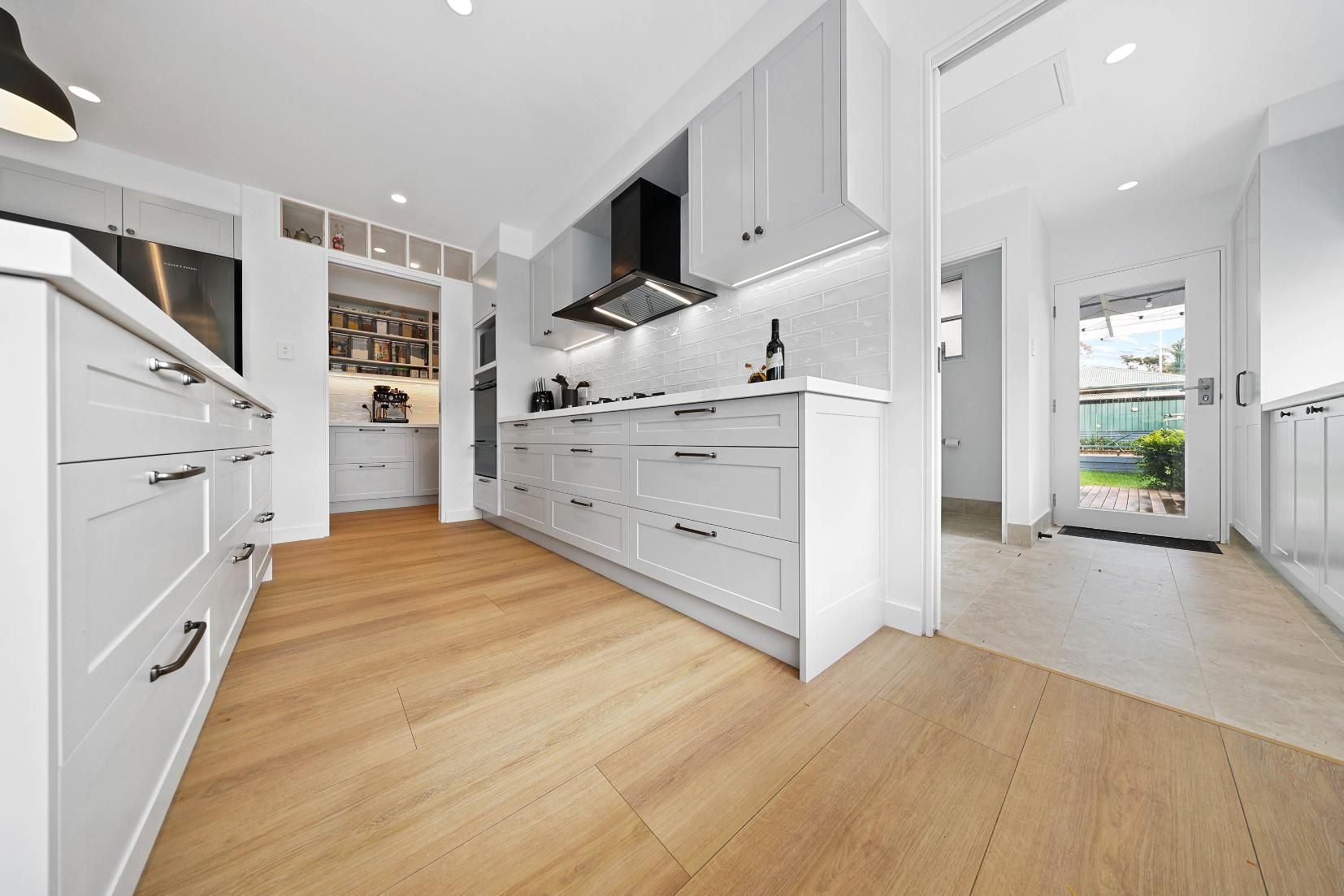 Light-Filled Kitchen with White Cabinetry, Light Wood Floors, Black Range Hood, and A View Into a Hallway — Above & Beyond Interiors Custom Joinery in Yowie Bay, NSW
