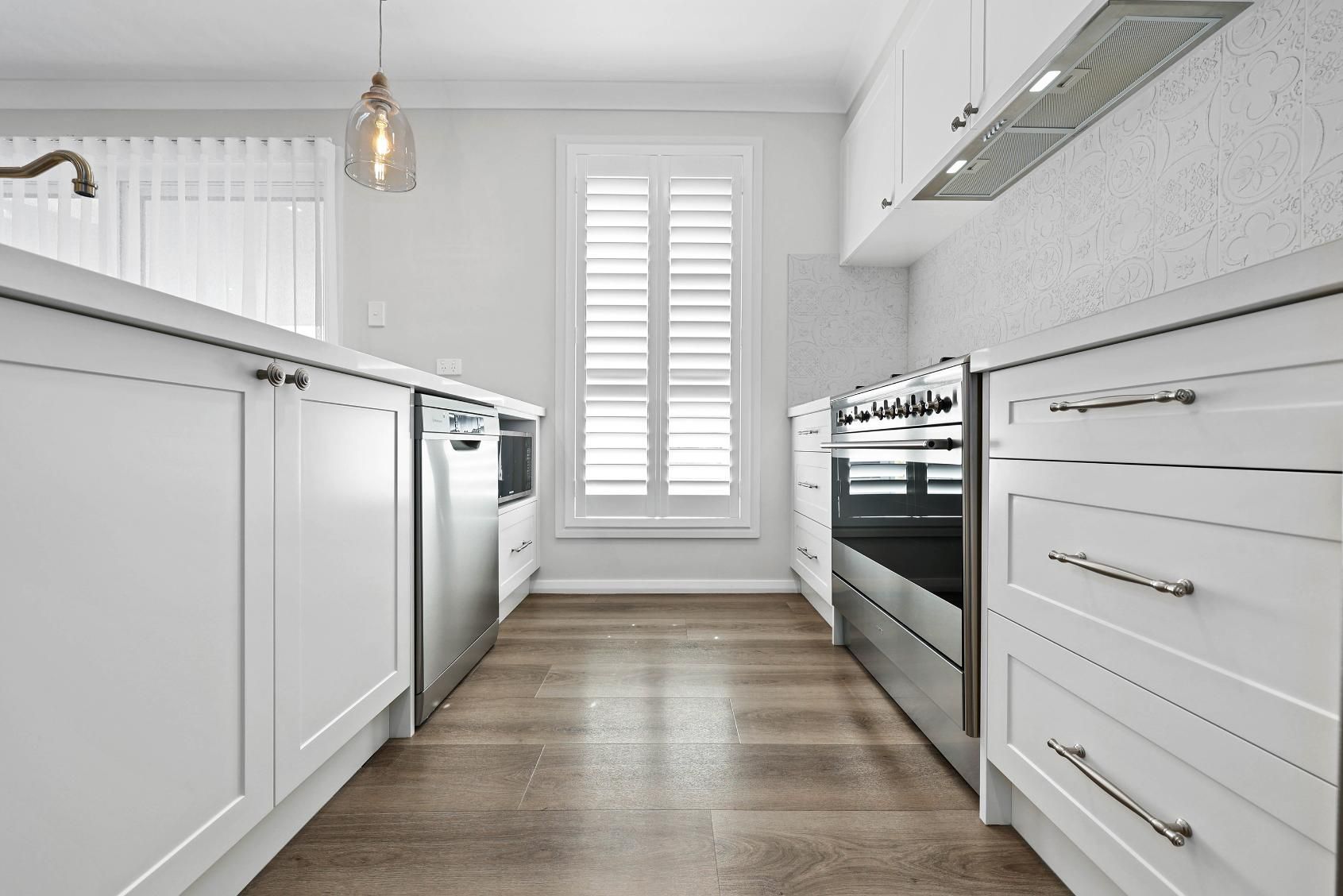 White Kitchen with Stainless Steel Appliances, Wood Floors, and A Window with Shutters — Above & Beyond Interiors Custom Joinery in Thirlmere, NSW