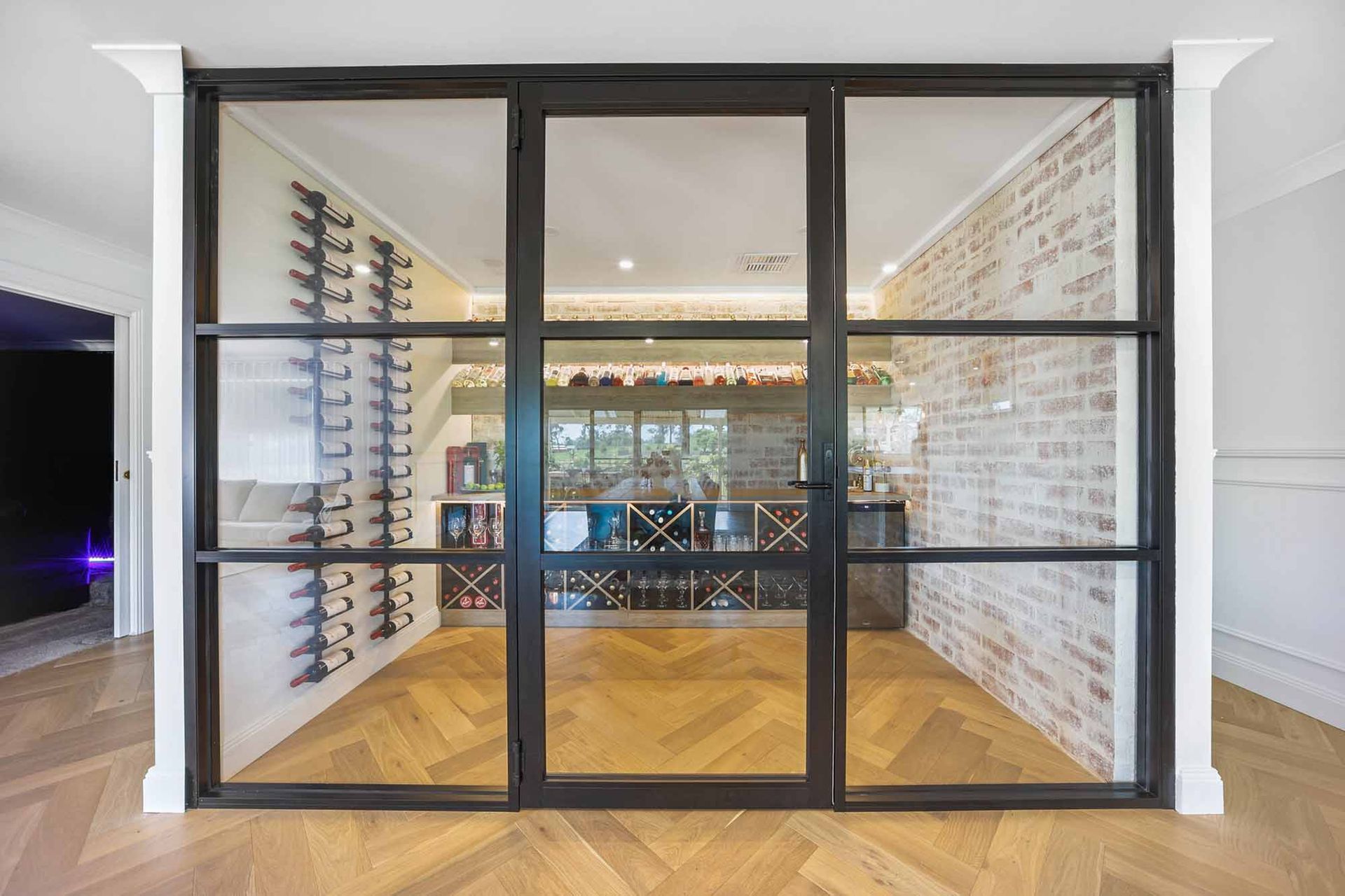 Black-framed glass doors reveal a wine room with brick wall, wine rack, and wooden floor — Above & Beyond Interiors Custom Joinery in Bellambi, NSW