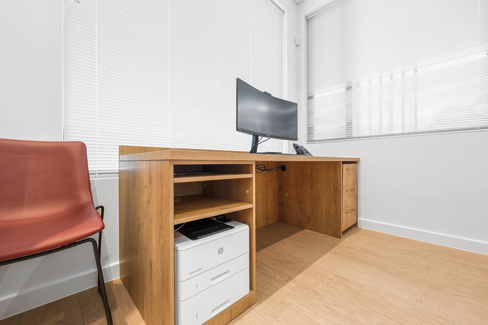 Office Desk with Computer Monitor Near a Window with Blinds. Red Chair to The Left — Above & Beyond Interiors Custom Joinery in Medical Centre, NSW