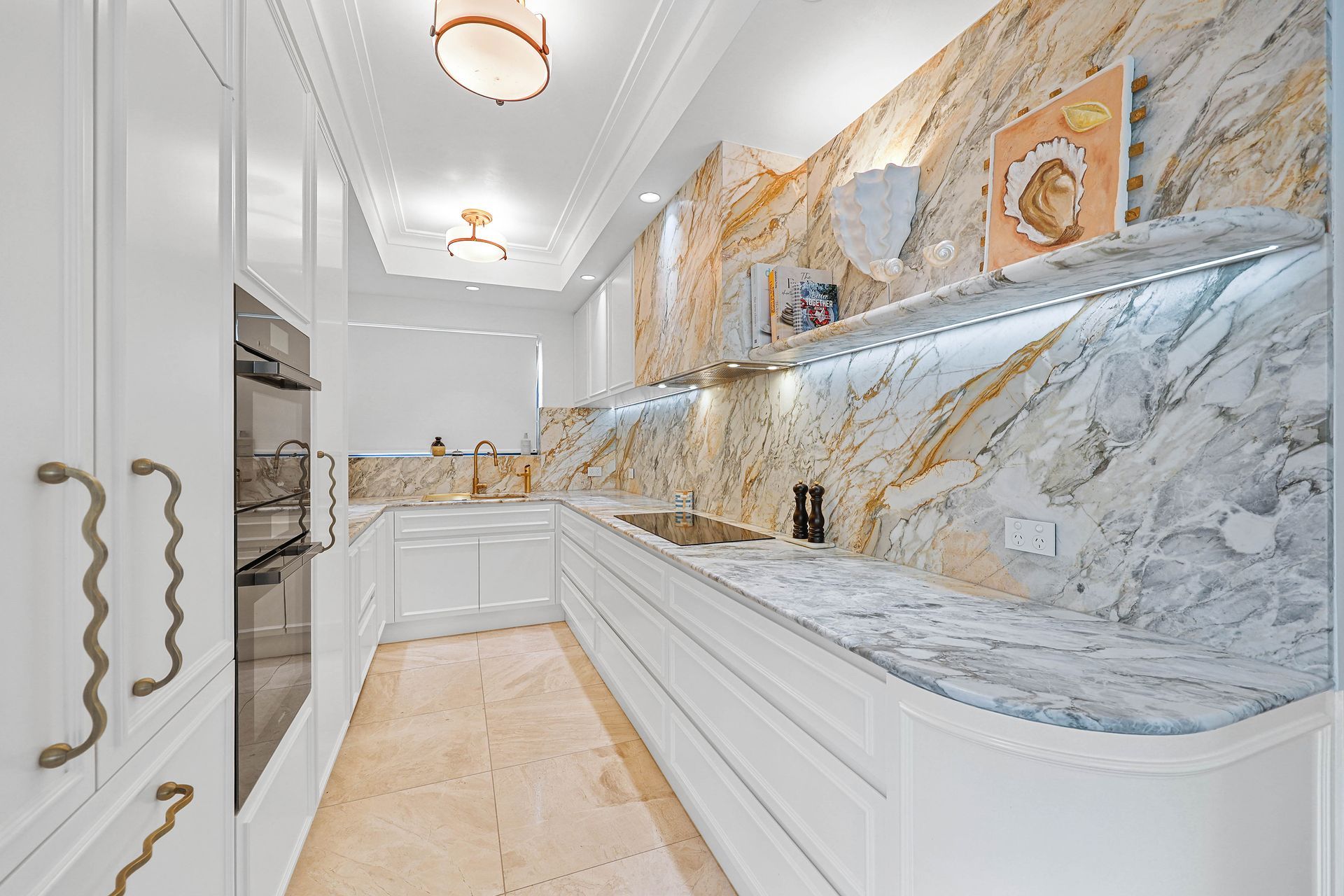 White and gold kitchen with marble countertops and backsplash, a small shelf, and appliances — Above & Beyond Interiors in Smeaton Grange, NSW