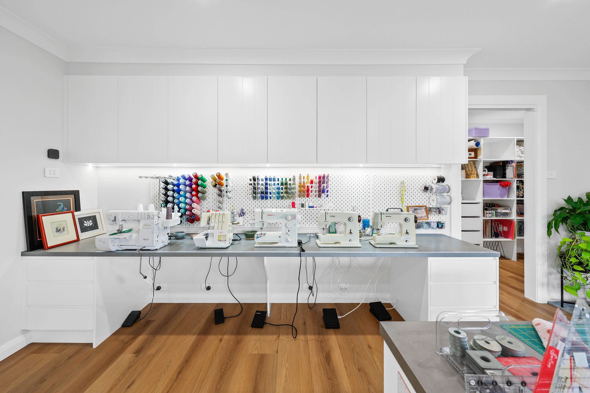 Sewing room with multiple machines on a long countertop, white cabinets, pegboard with spools, and wooden floors — Above & Beyond Interiors Custom Joinery in Bellambi, NSW