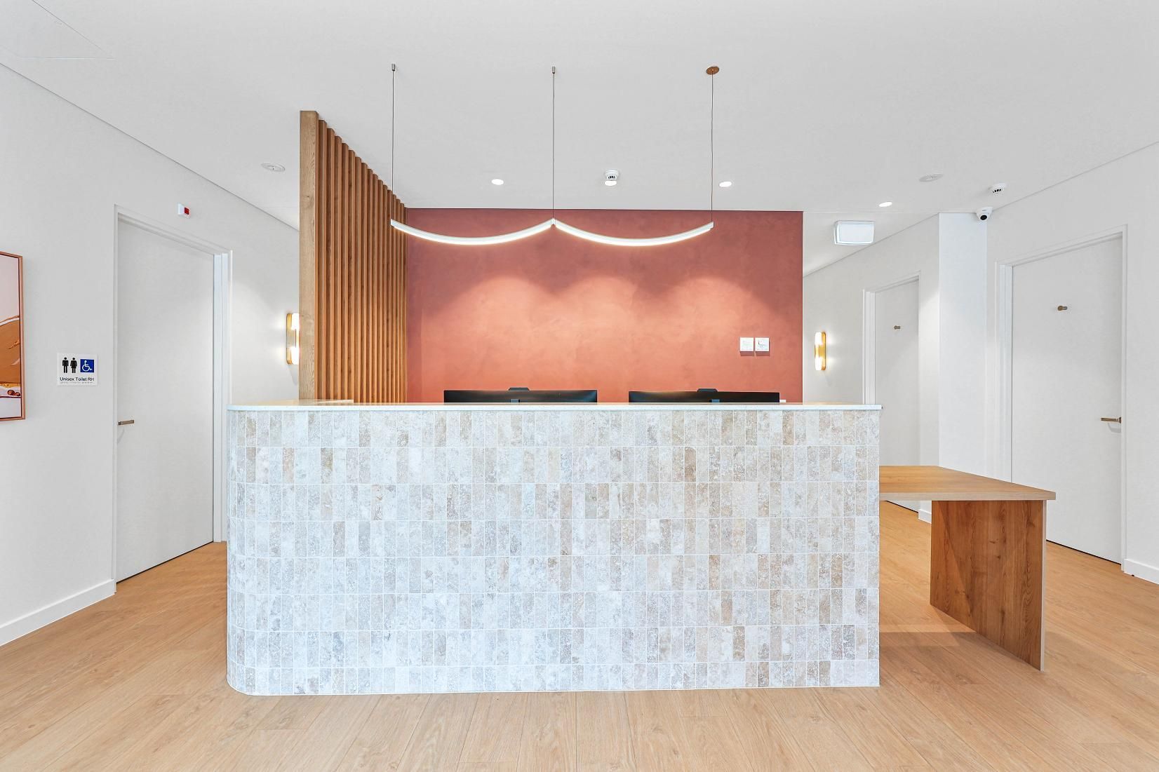 Reception Desk with Pink Wall, Wood Panel, and Light Fixture. Wooden Floor and White Doors — Above & Beyond Interiors Custom Joinery in Medical Centre, NSW
