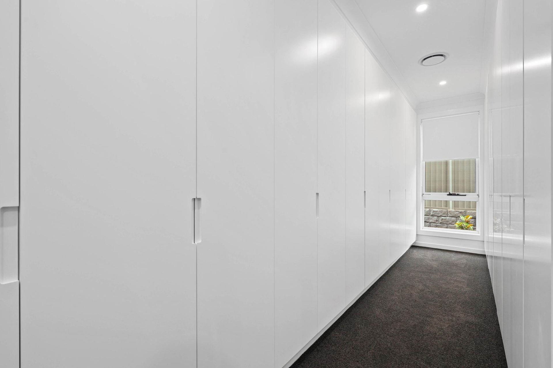 White Built-In Wardrobes Line a Hallway with Dark Carpet, Leading to A Window with A View — Above & Beyond Interiors Custom Joinery in Currans Hill, NSW