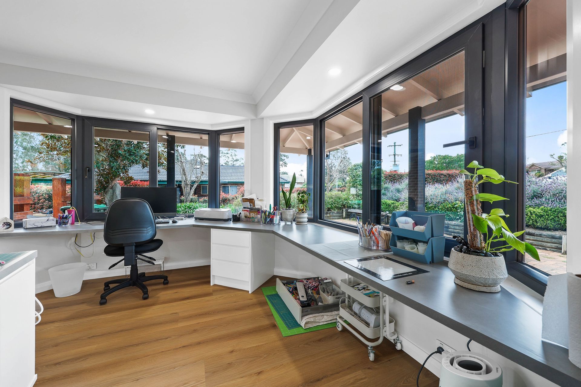 Bright home office with wraparound windows, desk, office chair, and plants, overlooking a yard — Above & Beyond Interiors Custom Joinery in Bellambi, NSW