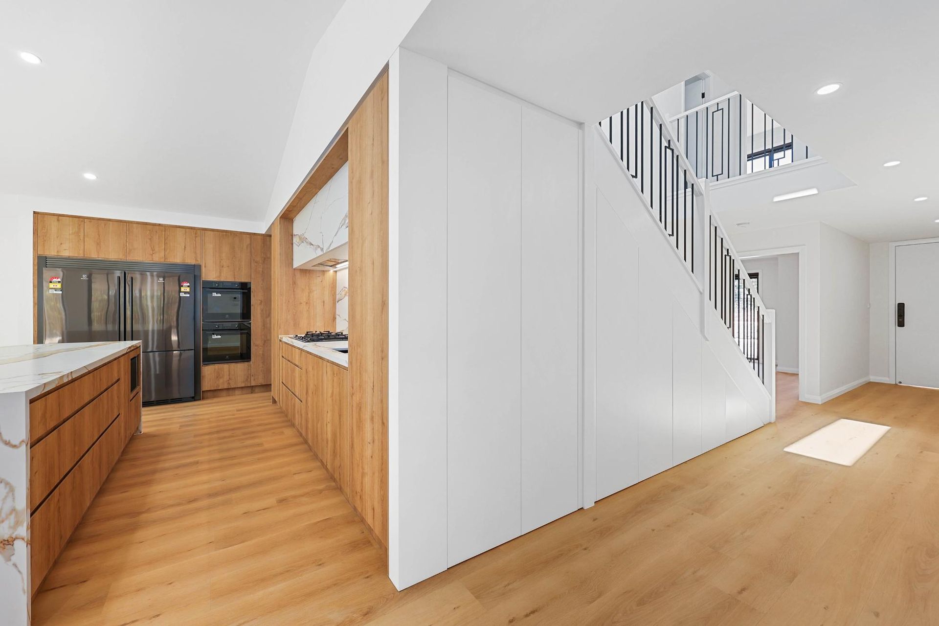 Modern Kitchen and Hallway with Wood Cabinets, Marble Island, and Staircase with Black Railing — Above & Beyond Interiors Custom Joinery in Abbortsbury, NSW