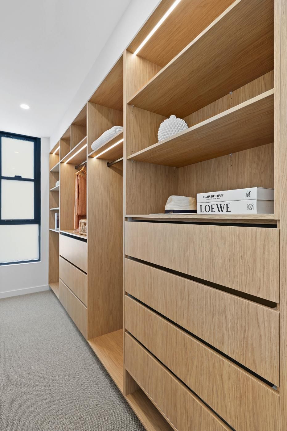 Wooden Walk-In Closet with Shelves, Drawers, and Hanging Space. Light-Colored Wood with A Neutral Carpet Floor — Above & Beyond Interiors Custom Joinery in Thirroul, NSW