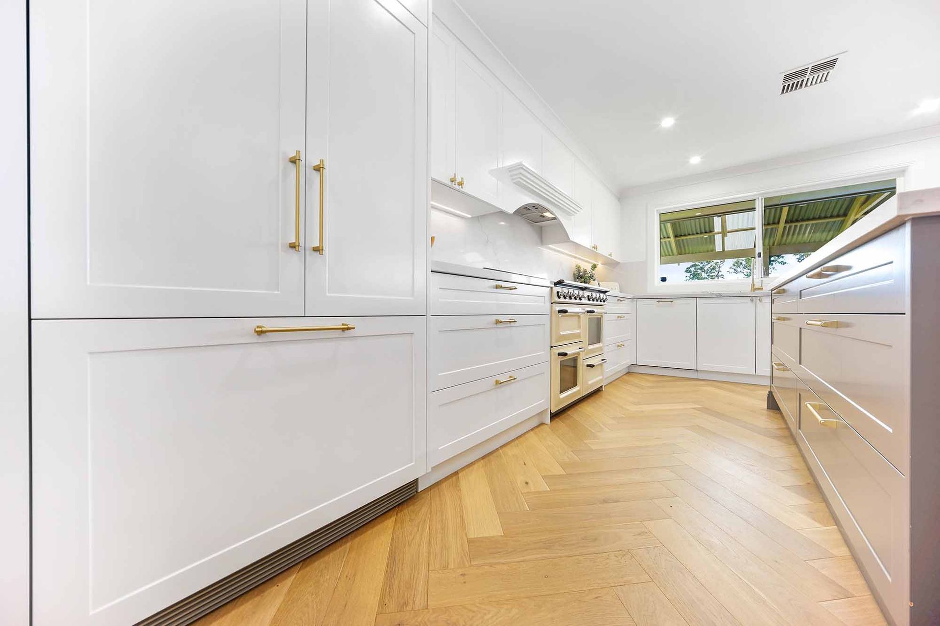 White kitchen with herringbone wood floor, cabinets with gold handles, and a window — Above & Beyond Interiors Custom Joinery in Bellambi, NSW