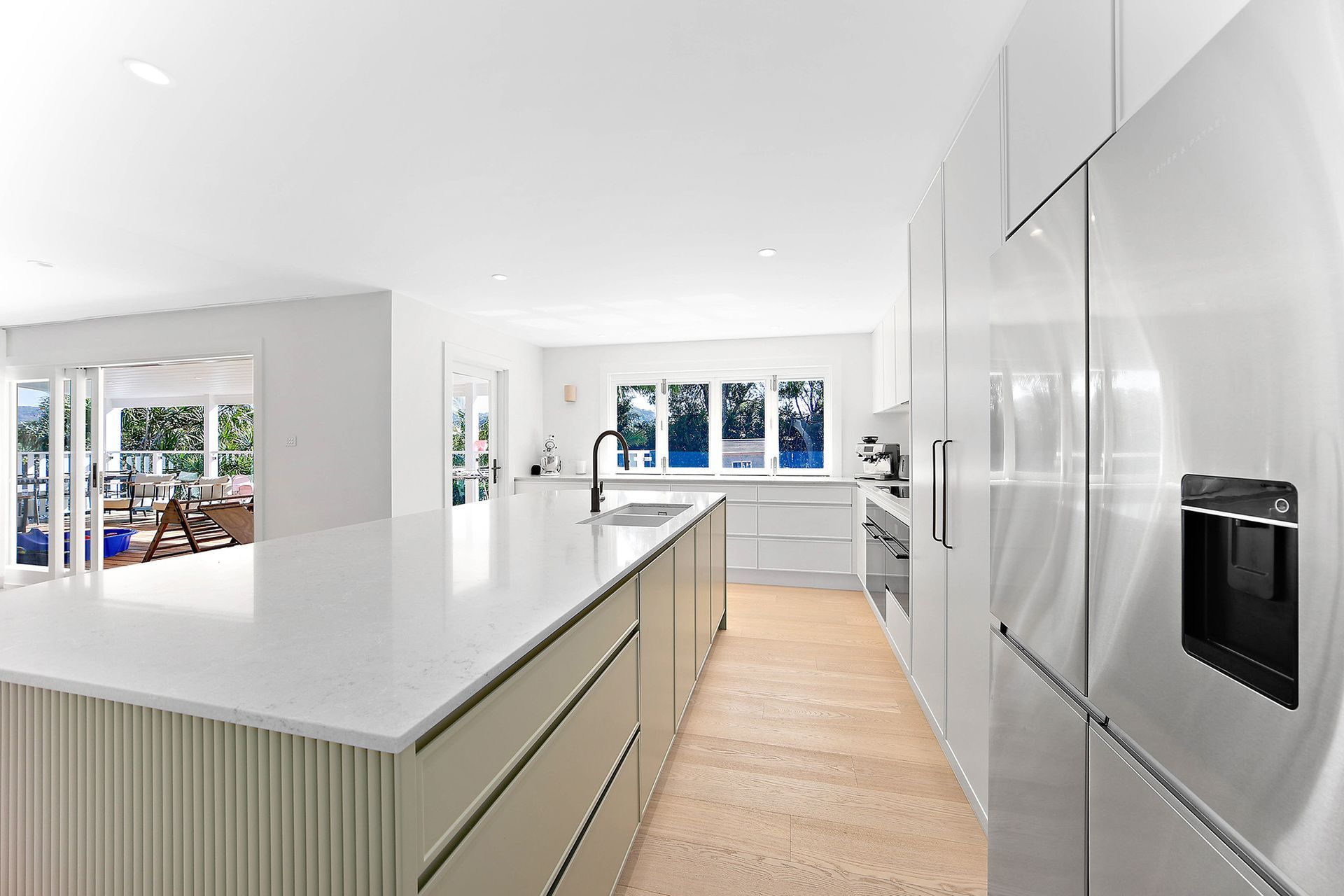 Modern white kitchen with large island and stainless steel refrigerator, overlooking an outdoor space — Above & Beyond Interiors Custom Joinery in Bellambi, NSW