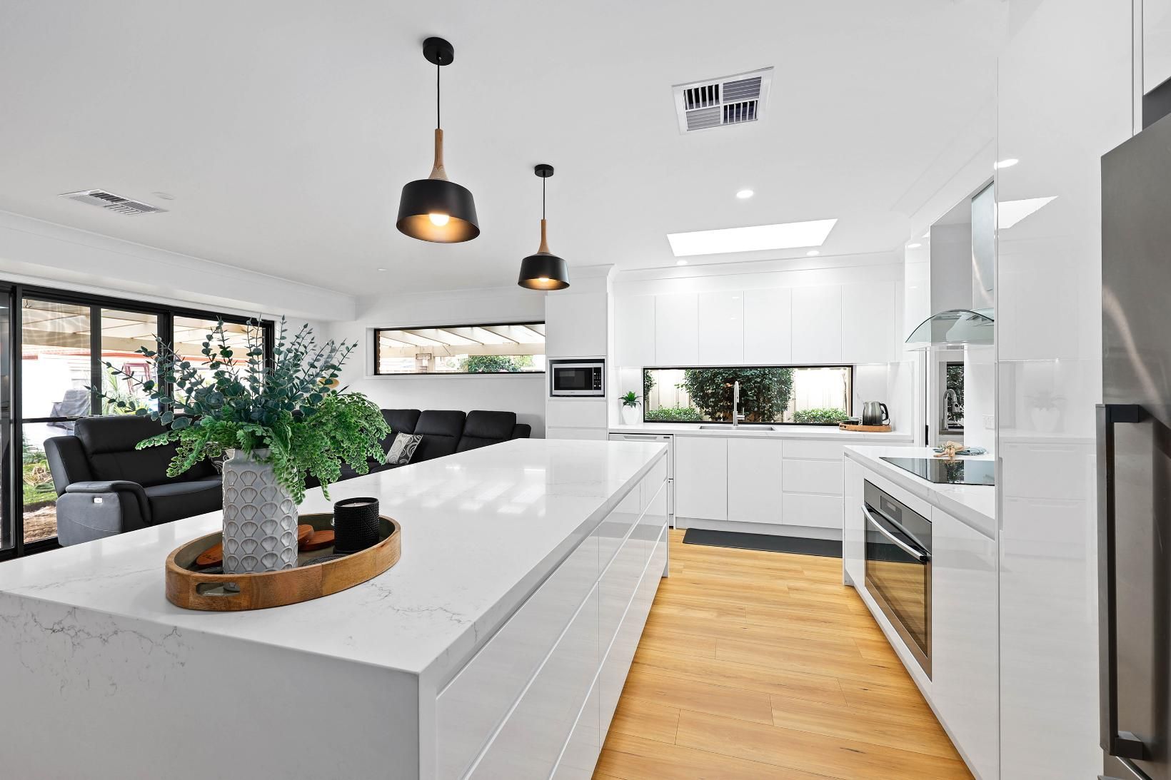 Modern White Kitchen with Large Island, Light Wood Floor, Open to Living Area with Black Sofa — Above & Beyond Interiors Custom Joinery in Wattle Grove, NSW