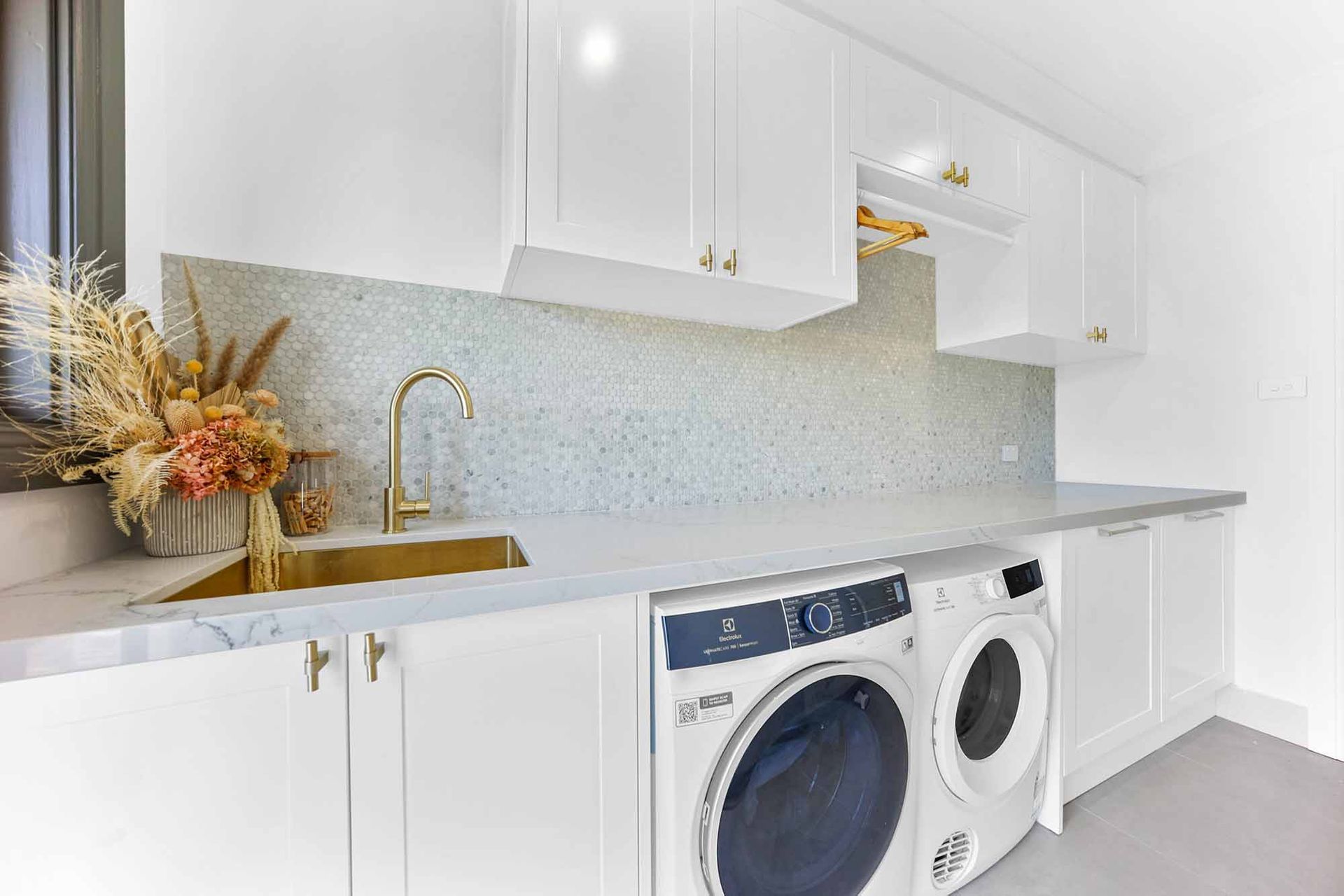 White laundry room with a marble countertop, gold faucet, and stacked washer/dryer — Above & Beyond Interiors Custom Joinery in Bellambi, NSW