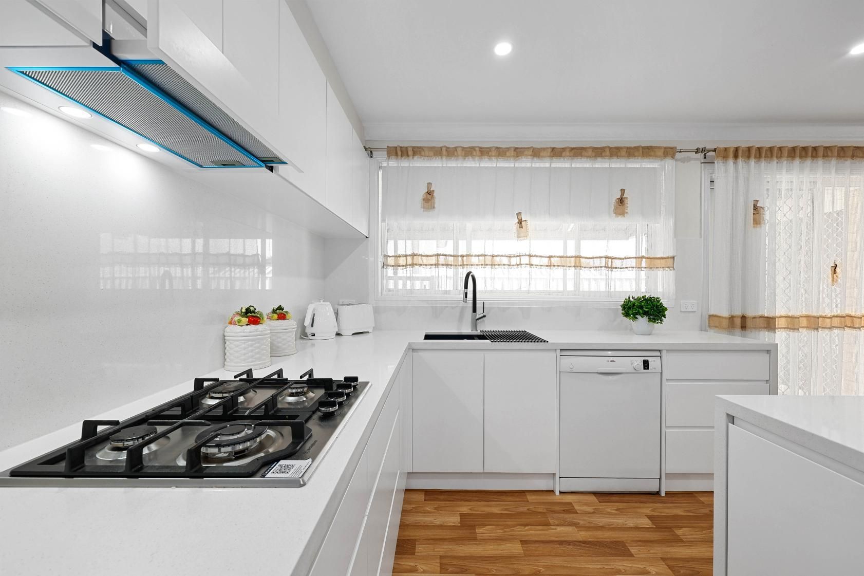 White Kitchen with Gas Stove, Sink, and Window with Light-Colored Curtains and Wooden Floor — Above & Beyond Interiors Custom Joinery in Narellan Vale, NSW