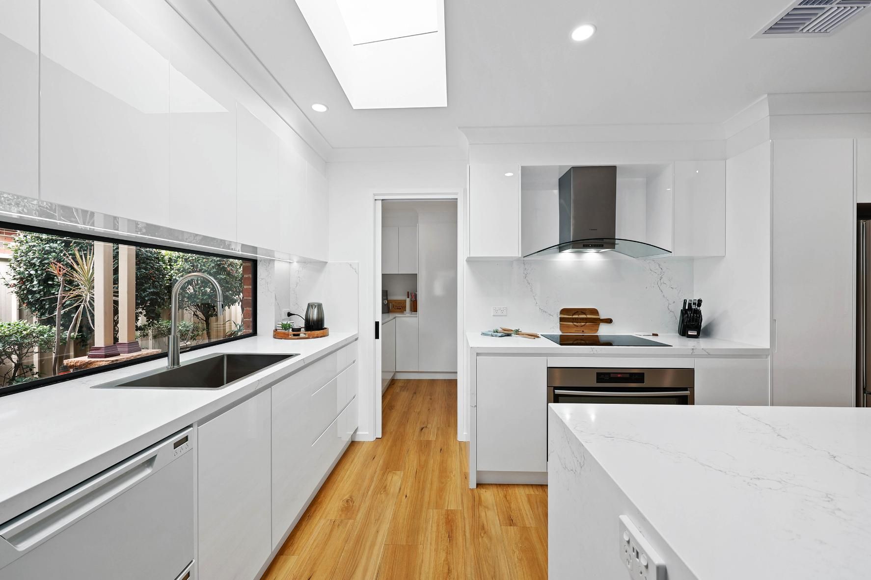 White Modern Kitchen with A Large Window, Stainless Steel Appliances, and Wooden Floor — Above & Beyond Interiors Custom Joinery in Wattle Grove, NSW
