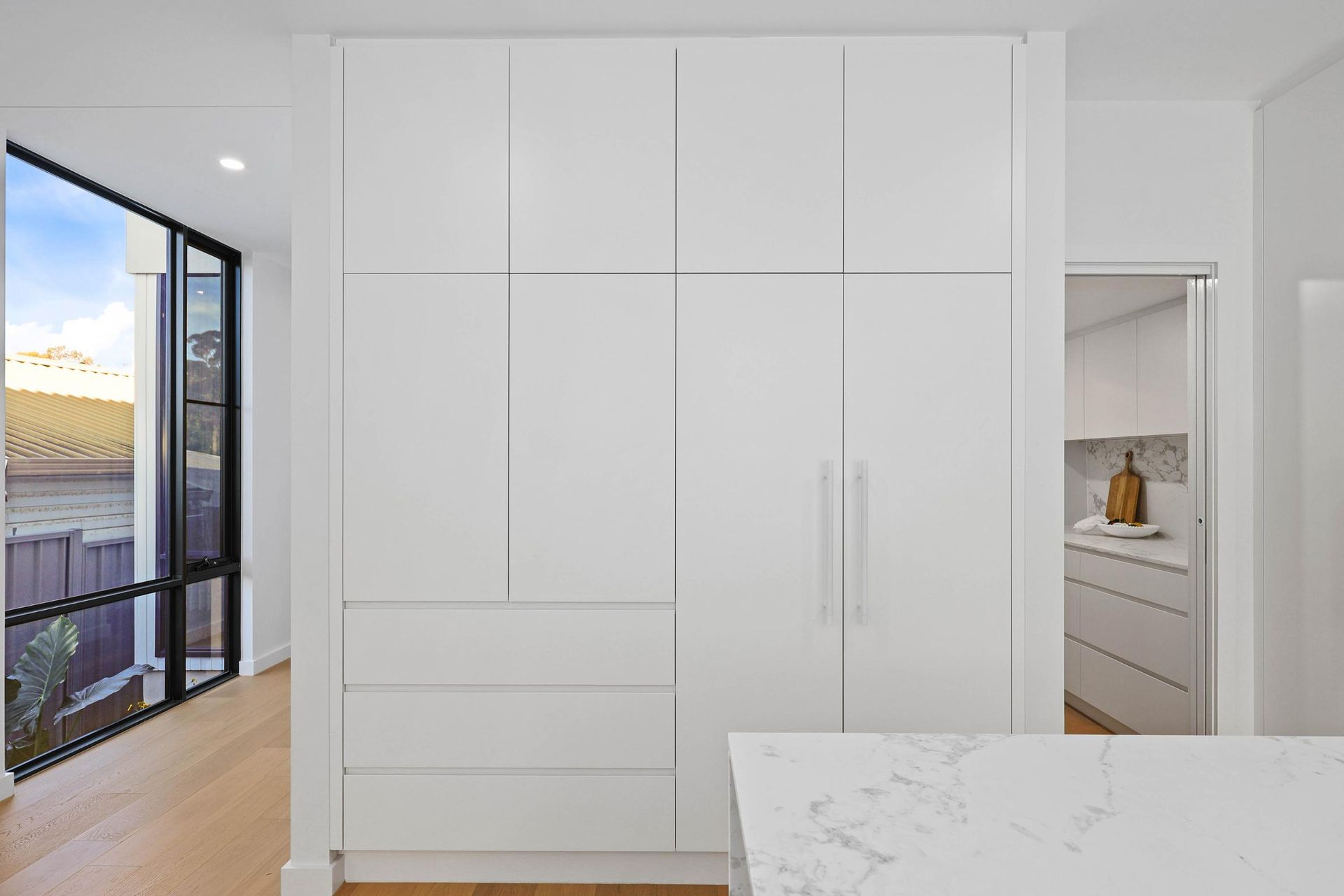 White Built-In Cabinets with Drawers and Long Handles, Beside a Window and Doorway, with A Marble Counter — Above & Beyond Interiors Custom Joinery in Thirroul, NSW