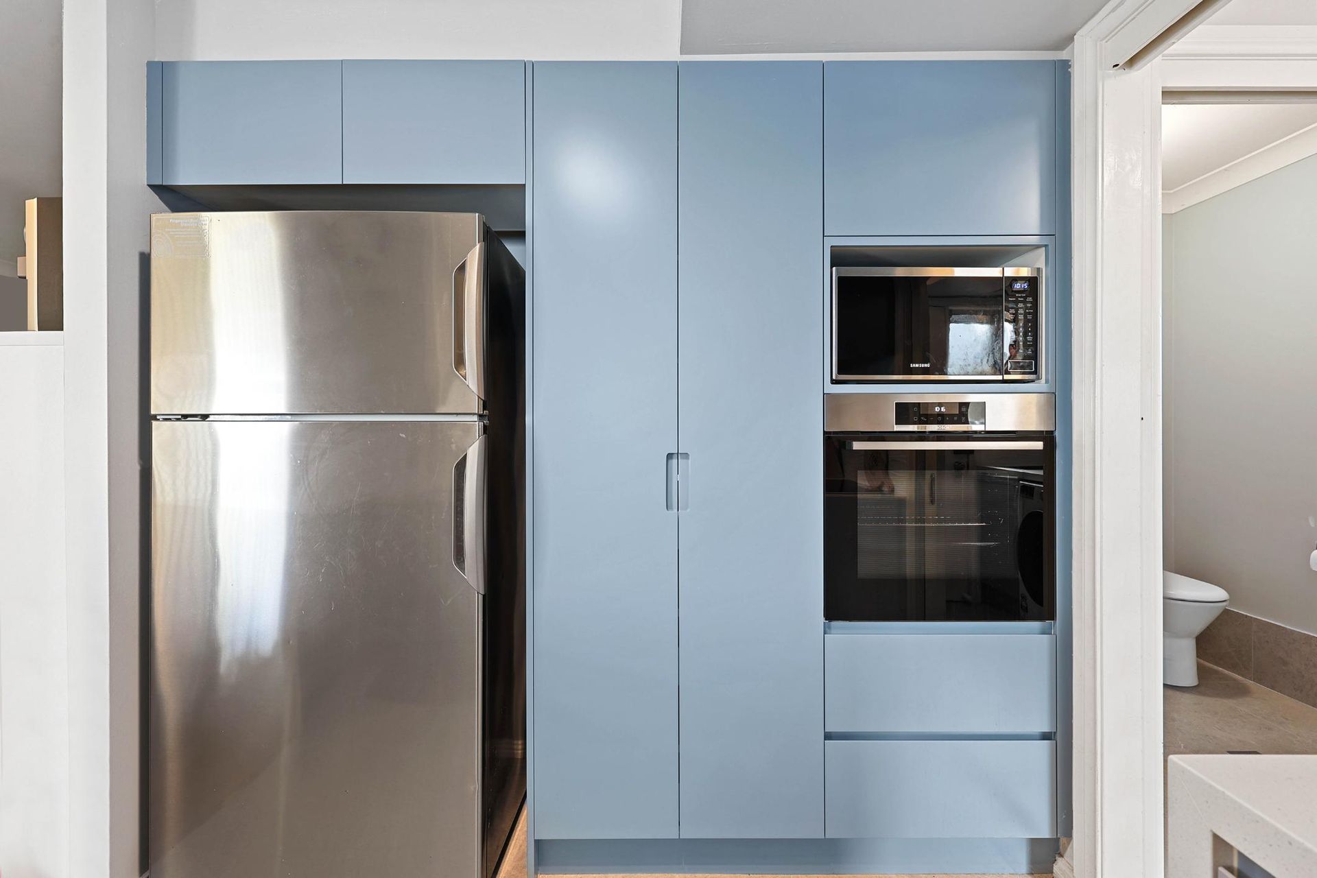 Stainless Steel Refrigerator Next to Light Blue Kitchen Cabinetry with Microwave and Oven — Above & Beyond Interiors Custom Joinery in Macquarie Fields, NSW