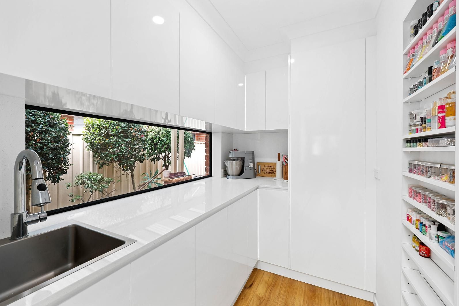 Modern White Kitchen with A Long Window Overlooking Greenery and A Stainless Steel Sink — Above & Beyond Interiors Custom Joinery in Wattle Grove, NSW