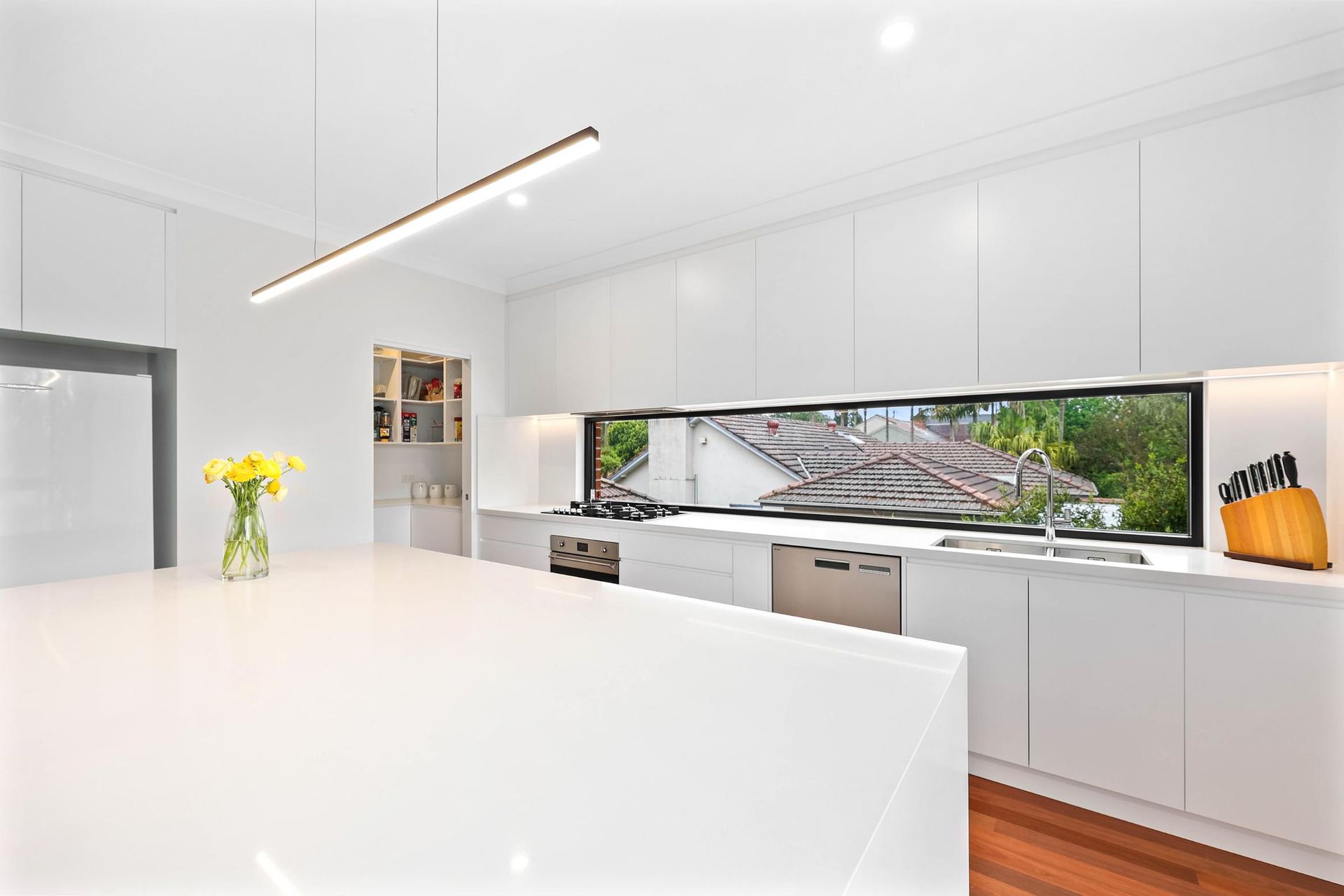 Modern White Kitchen with Large Island, Long Window Showing Rooftops, and A Linear Pendant Light — Above & Beyond Interiors Custom Joinery in Killara, NSW