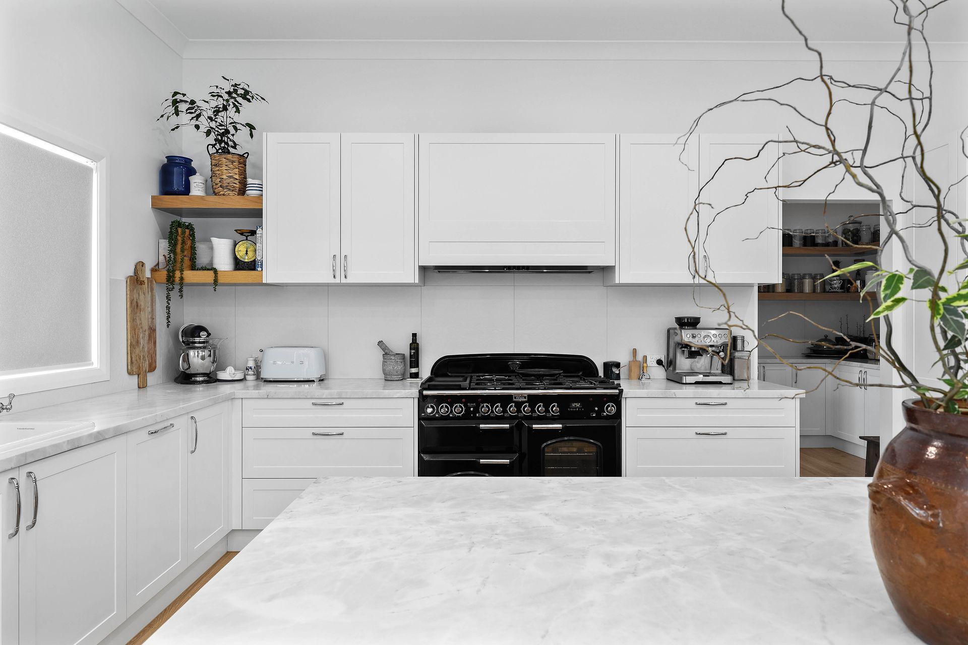 White kitchen with black range, marble countertops, and rustic vase with branches — Above & Beyond Interiors Custom Joinery in Bradbury, NSW
