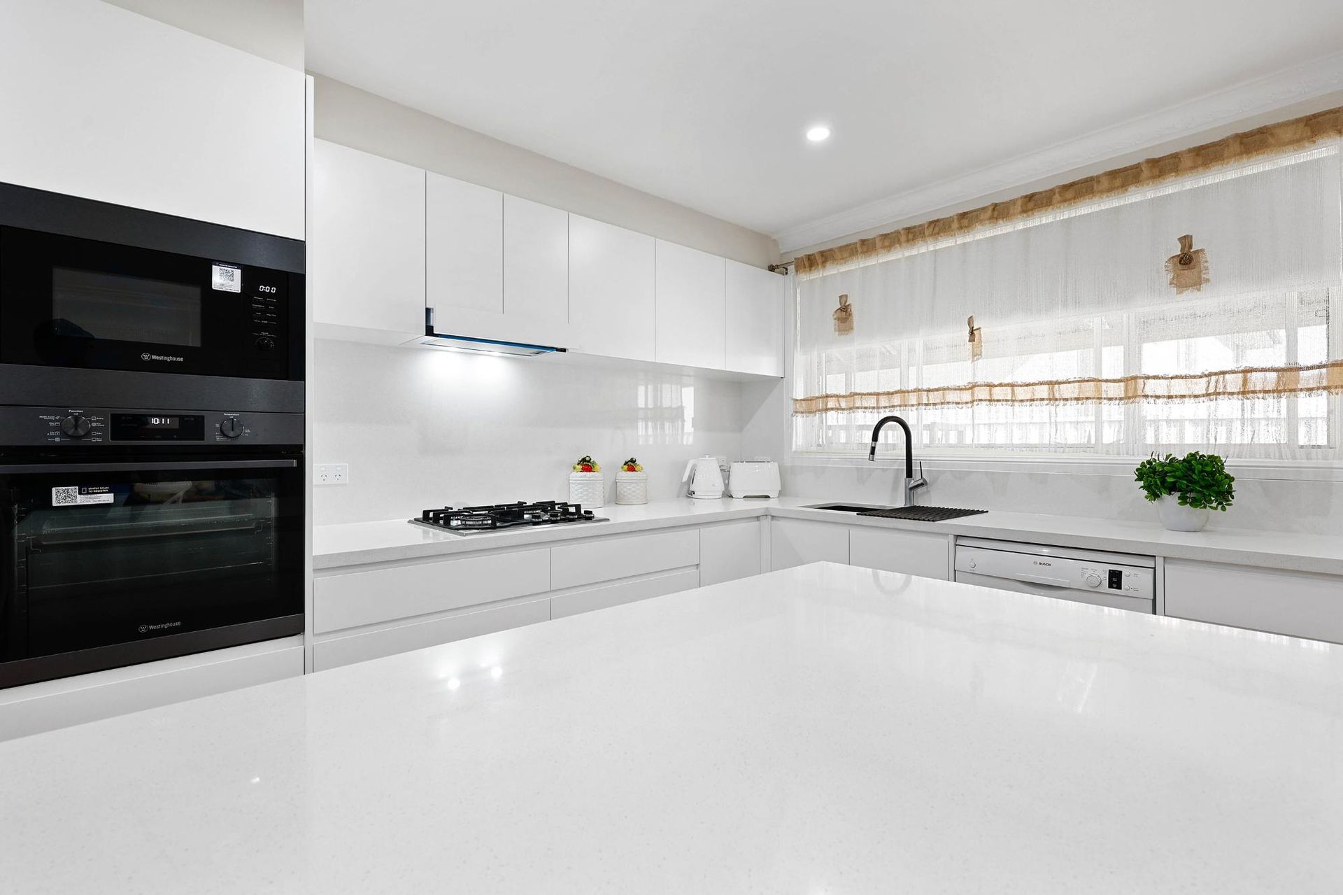 Modern, White Kitchen with Black Appliances, White Countertops, and A Window with Sheer Curtains — Above & Beyond Interiors Custom Joinery in Narellan Vale, NSW