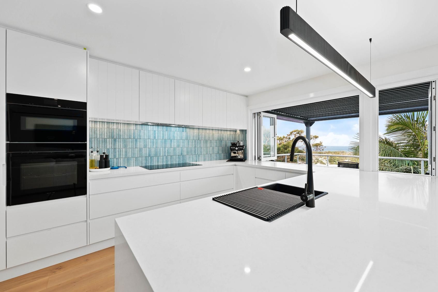 Modern White Kitchen with Blue Backsplash, Black Appliances, and An Island with A Sink Overlooking an Outdoor View — Above & Beyond Interiors Custom Joinery in North Curl Curl, NSW
