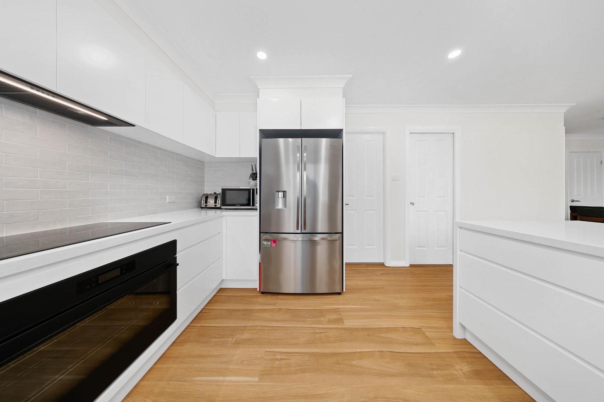 Modern White Kitchen with Stainless Steel Refrigerator, Light Wood Floors, and White Cabinetry — Above & Beyond Interiors Custom Joinery in Mount Anna, NSW